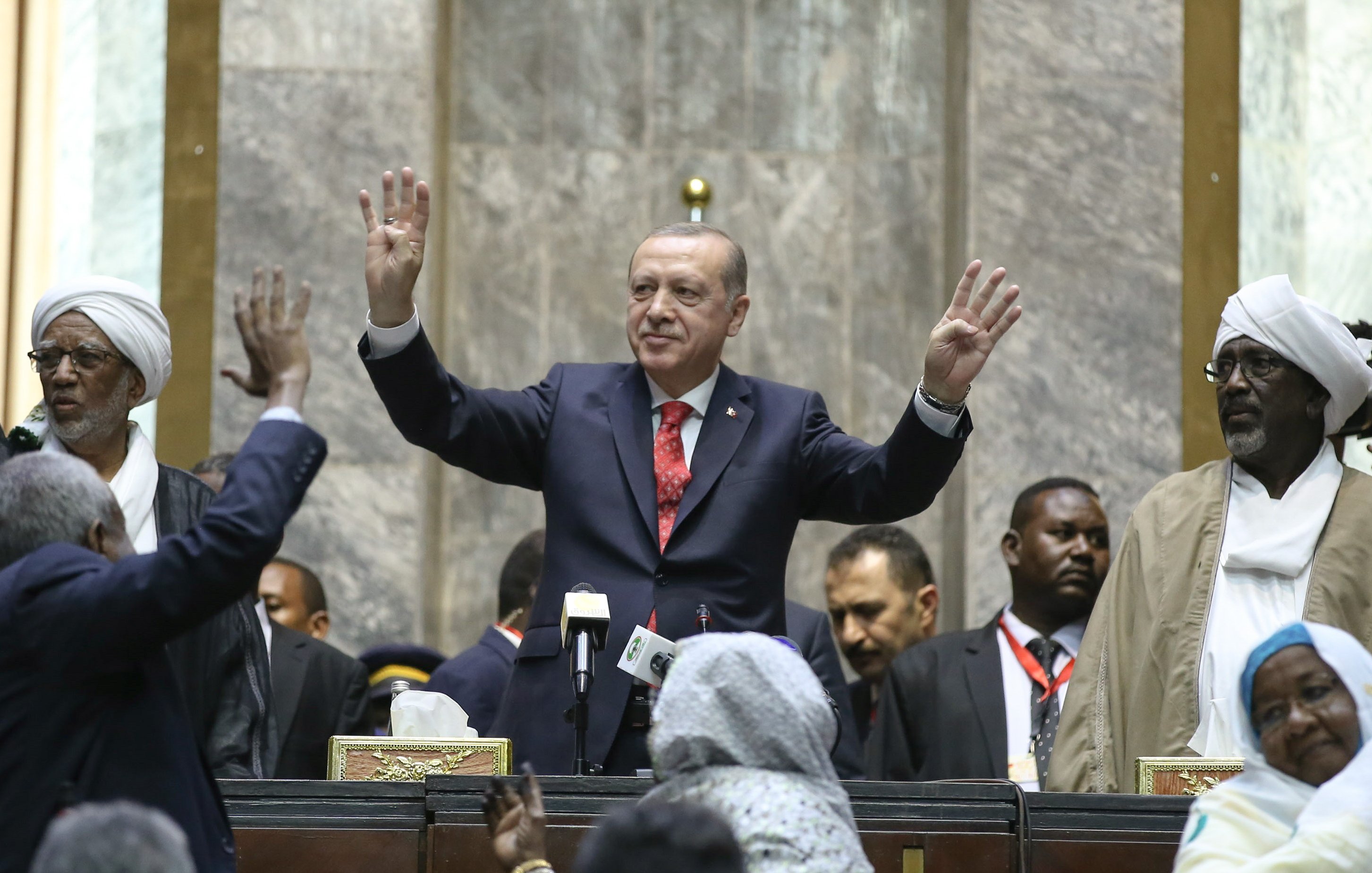 تظهر الصورة تجمعًا سياسيًا حيث يقف شخصية معروفة في المنتصف، مرتديًا بدلة رسمية، ويقوم بإشارة بيديه. خلفه يظهر عدد من الأشخاص، بعضهم يبدو متحمسًا ويرفع أيديه، بينما يوجد آخرون يقفون بجانبه. المكان يبدو رسميًا، مع وجود خلفية مكونة من جدران حجرية وأعلام. الأجواء تعكس حماس الحضور.