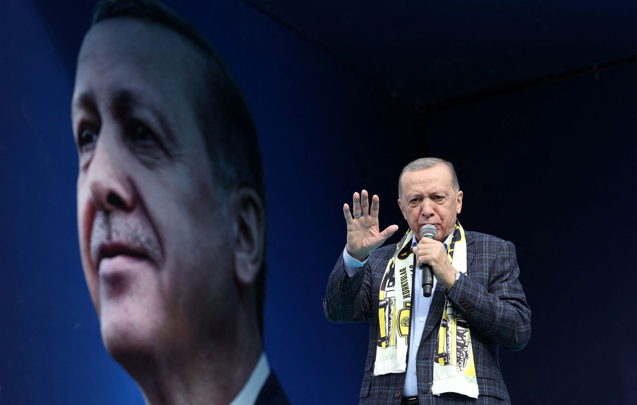 L'image montre un homme sur une scène, tenant un microphone et s'adressant à une foule. Il porte une écharpe aux couleurs d'une équipe de football et semble faire un discours passionné. En arrière-plan, on peut voir un grand portrait de lui, qui renforce sa présence sur scène. La scène est ornée d'un fond bleu, mettant en avant son image.