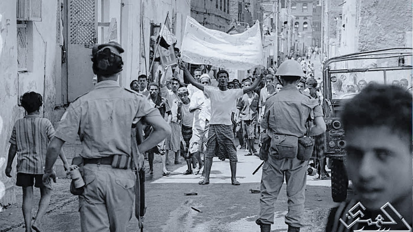 L'image montre une scène de rue dans un contexte de manifestation ou de rassemblement. On peut voir des groupes de personnes, certains brandissant des drapeaux ou des pancartes, exprimant visiblement leur émotion ou leur revendication. Des policiers réglementent la situation, et l'atmosphère semble tendue. L'environnement urbain est composé de bâtiments simples, typiques d'une ville. Les vêtements des gens indiquent peut-être une époque passée, soulignant le contexte historique de la scène.