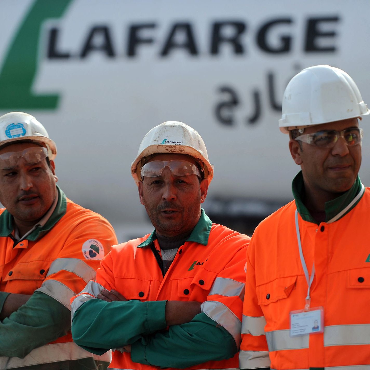 تظهر الصورة ثلاثة رجال يرتدون ملابس العمل البرتقالية، ويضعون خوذات بيضاء على رؤوسهم. يبدو أنهم يعملون في موقع بناء أو مصنع، حيث خلفهم تظهر شاحنة أو معدات تحمل اسم "لافارج". يعبر تعبيرهم عن الجدية والتركيز في العمل، بينما يقفون معًا.