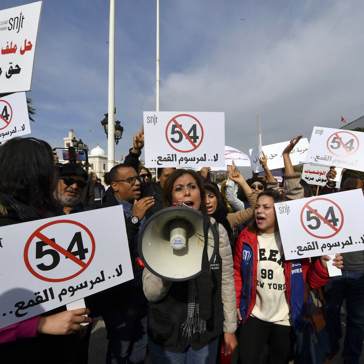 The image depicts a protest scene with a group of people holding up signs and banners. Some demonstrators are prominently featuring a sign with the number "54" crossed out, suggesting opposition to a specific law or regulation. One woman is speaking into a megaphone, leading the crowd in their protest. The atmosphere appears energetic and determined, with a focus on raising awareness or voicing their concerns regarding a particular issue.