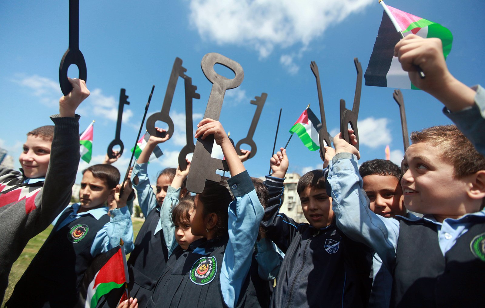 L'image montre un groupe d'enfants, qui semblent être en train de célébrer ou de manifester. Ils tiennent des grandes clés en carton et des drapeaux palestiniens. Les enfants, vêtus de uniformes scolaires, sourient et lèvent les clés vers le ciel, symbolisant un message de revendication ou d'espoir. Le ciel est bleu avec quelques nuages, ce qui ajoute à l'ambiance de la scène.