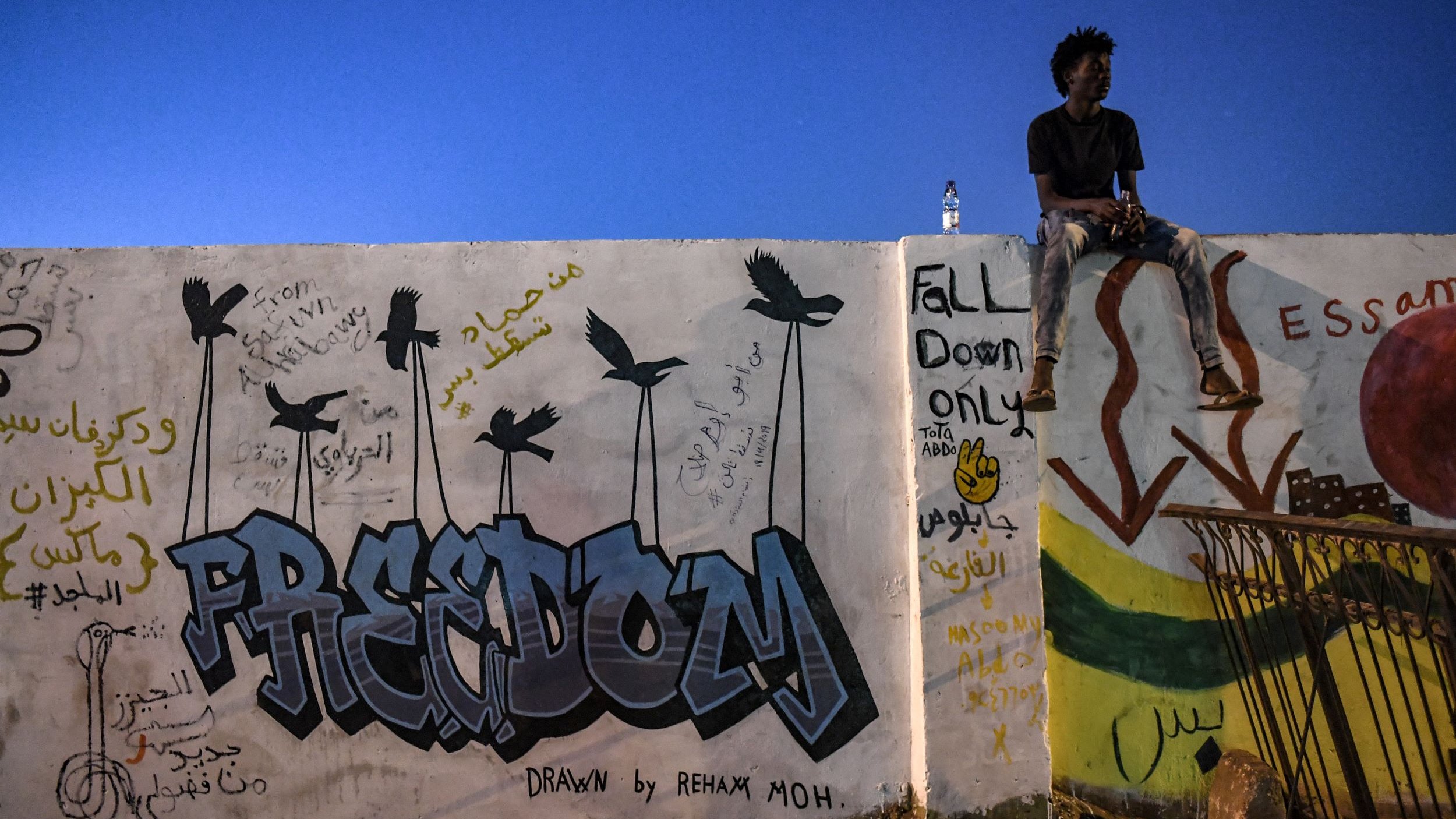 L'image montre un mur couvert de graffitis avec le mot "FREEDOM" en lettres colorées. À côté, des dessins d'oiseaux sont visibles. Un jeune homme est assis sur le bord du mur, regardant au loin, tandis qu'une bouteille d'eau est posée à côté de lui. Le ciel est d'un bleu profond, indiquant une prise de vue en soirée. Des inscriptions en arabe et des dessins supplémentaires peuvent être observés sur le mur.