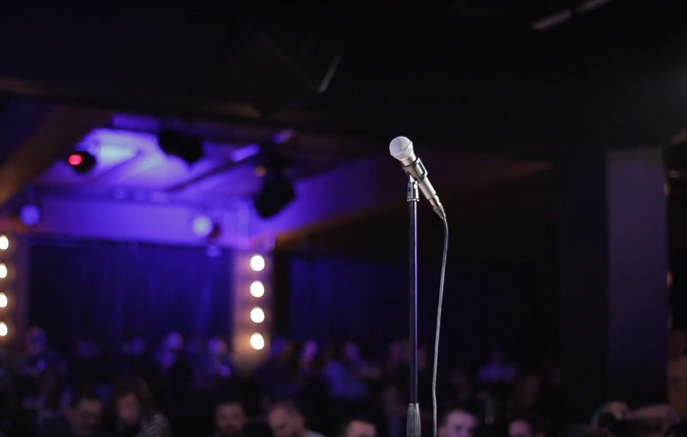 L'image montre un microphone sur un pied, au premier plan, sur une scène sombre. À l'arrière-plan, on aperçoit une audience floue et des lumières tamisées qui créent une ambiance de spectacle.