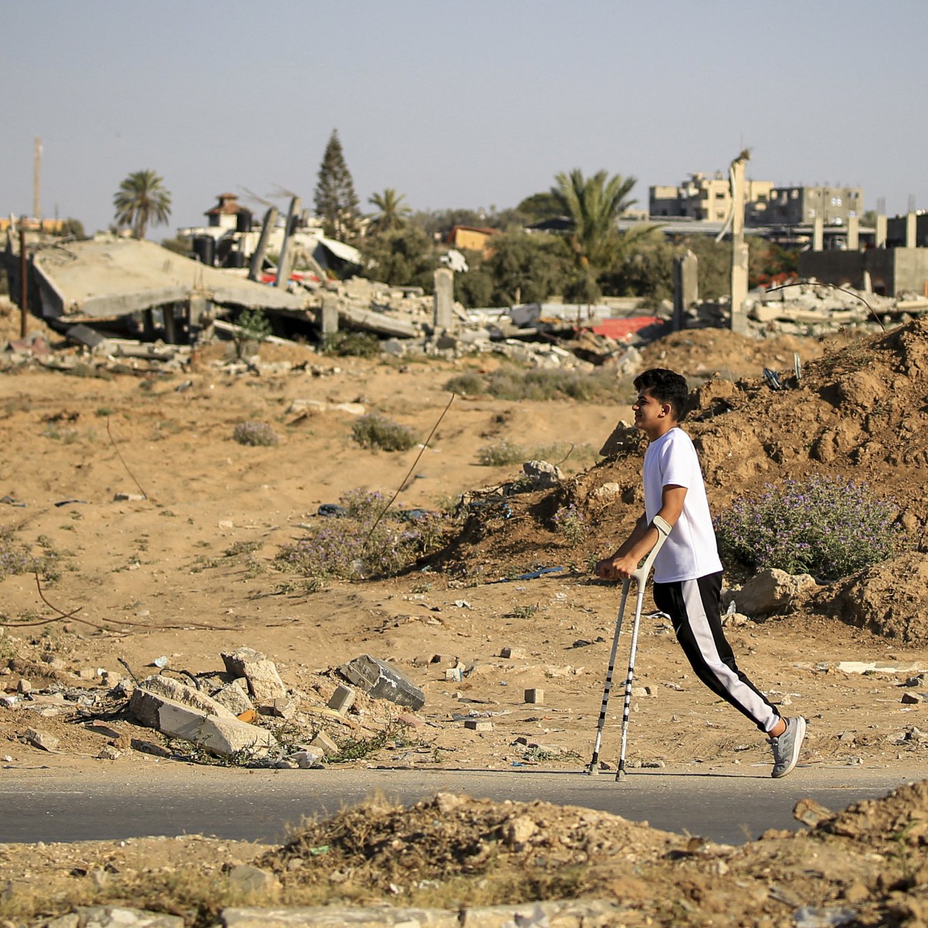 L'image montre un jeune garçon marchant avec des béquilles dans un paysage de destruction. En arrière-plan, on peut voir des bâtiments en ruines et des débris éparpillés, témoignant d'un environnement dévasté. Le sol est recouvert de terre et de petites herbes, créant un contraste avec les restes des structures. L'atmosphère semble sombre et tragique, soulignant la situation difficile dans laquelle se trouve l'enfant.