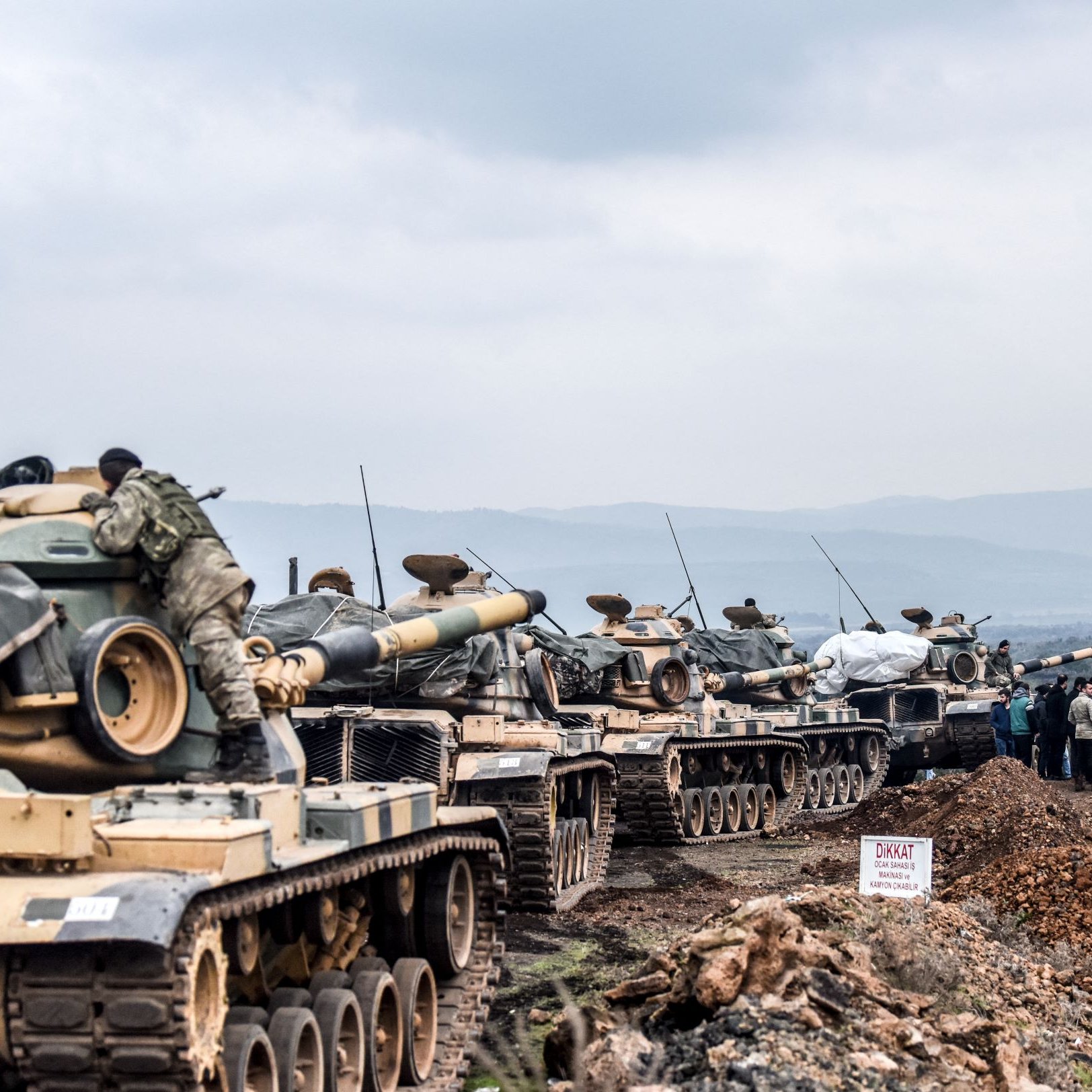 در این تصویر تعدادی تانک دیده می‌شود که در یک خط قرار گرفته‌اند. تانک‌ها به نظر می‌رسد به گونه‌ای آماده‌سازی شده‌اند و برخی از آن‌ها با پوشش‌هایی پوشیده شده‌اند. در پس‌زمینه، چند نفر در حال فعالیت هستند و دود از نقطه‌ای در دوردست به هوا برمی‌خیزد. مناظر طبیعی مانند تپه‌ها و آسمان ابری نیز در تصویر قابل مشاهده است. این صحنه می‌تواند به یک عملیات نظامی یا تمرینات نظامی اشاره داشته باشد.