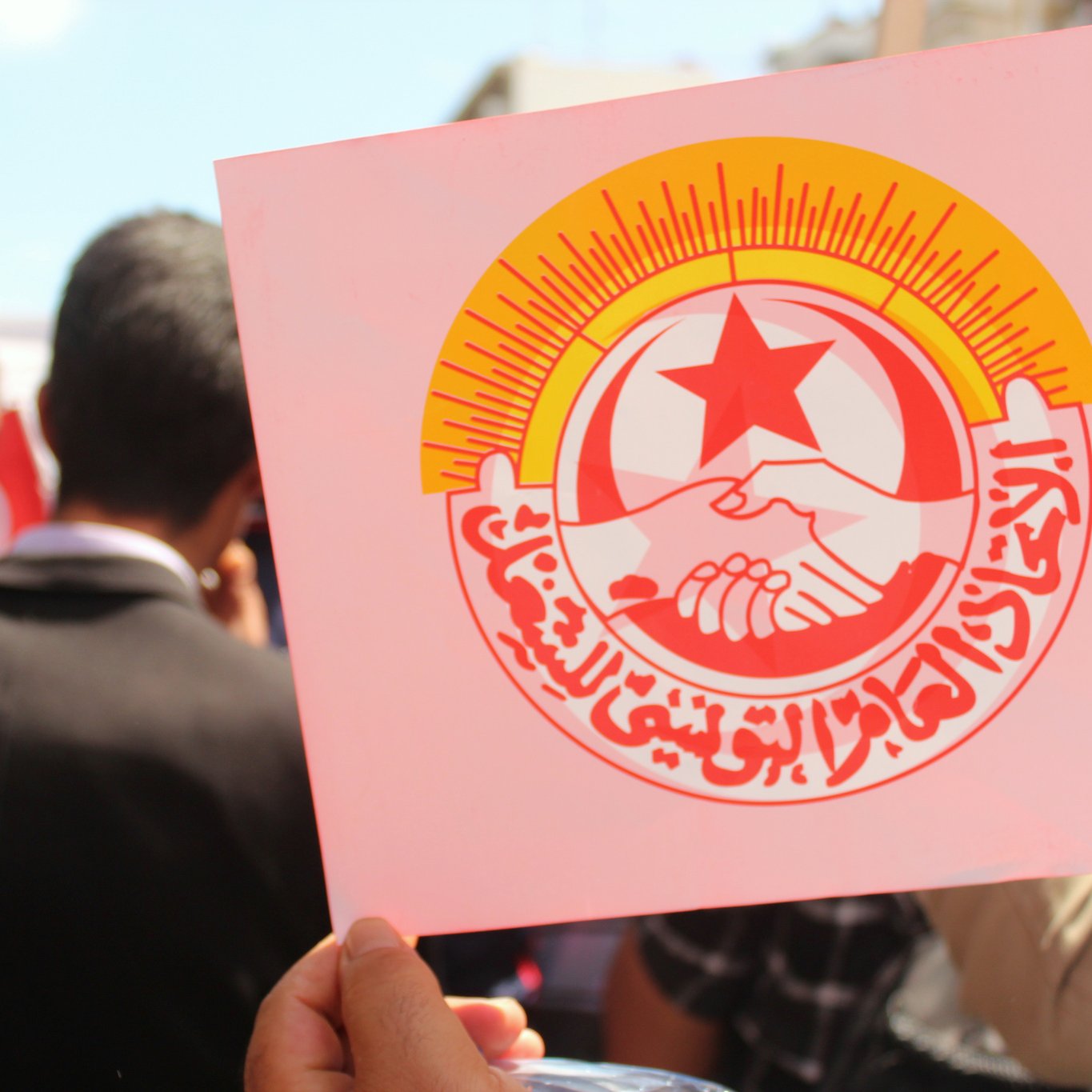 L'image montre une manifestation ou un rassemblement où une personne tient une pancarte avec un logo. Ce logo représente probablement une organisation syndicale ou politique, avec des éléments symboliques comme des mains qui se serrent et une étoile rouge. En arrière-plan, on peut apercevoir des gens qui semblent participer à l'événement, ainsi que des drapeaux. L'atmosphère semble être celle de solidarité et de mobilisation.