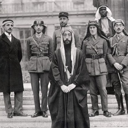 L'image montre un groupe de six hommes posant ensemble. Ils sont vêtus de différentes tenues militaires et traditionnelles. Au centre, un homme en robe traditionnelle, portant une tête couronnée, semble être le principal sujet. À sa gauche, un autre homme en uniforme militaire. Les autres sont également en uniformes variés, indiquant peut-être des origines et des rôles différents. L'arrière-plan montre une architecture qui semble historique. L'ensemble dégage une atmosphère de confluence culturelle et de collaboration.