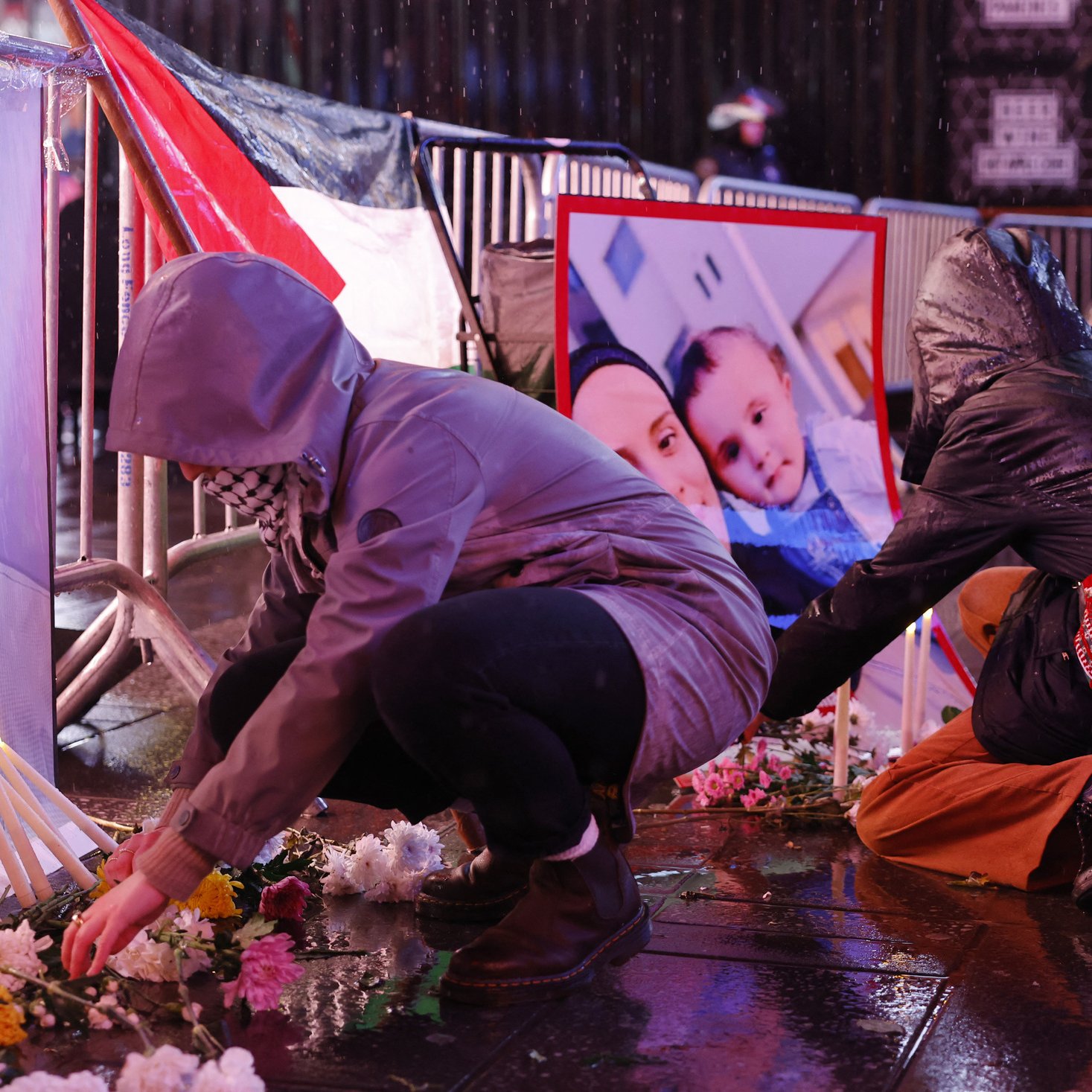 L'image montre des personnes en train de rendre hommage devant des portraits posés au sol. On peut voir des bougies allumées et des fleurs disposées autour des photos. Certaines personnes s'agenouillent ou se penchent pour arranger les fleurs. L'environnement semble humide, peut-être en raison de la pluie, et les participants portent des vêtements adaptés aux intempéries, comme des imperméables et des écharpes. L'atmosphère semble solennelle et empreinte de respect.