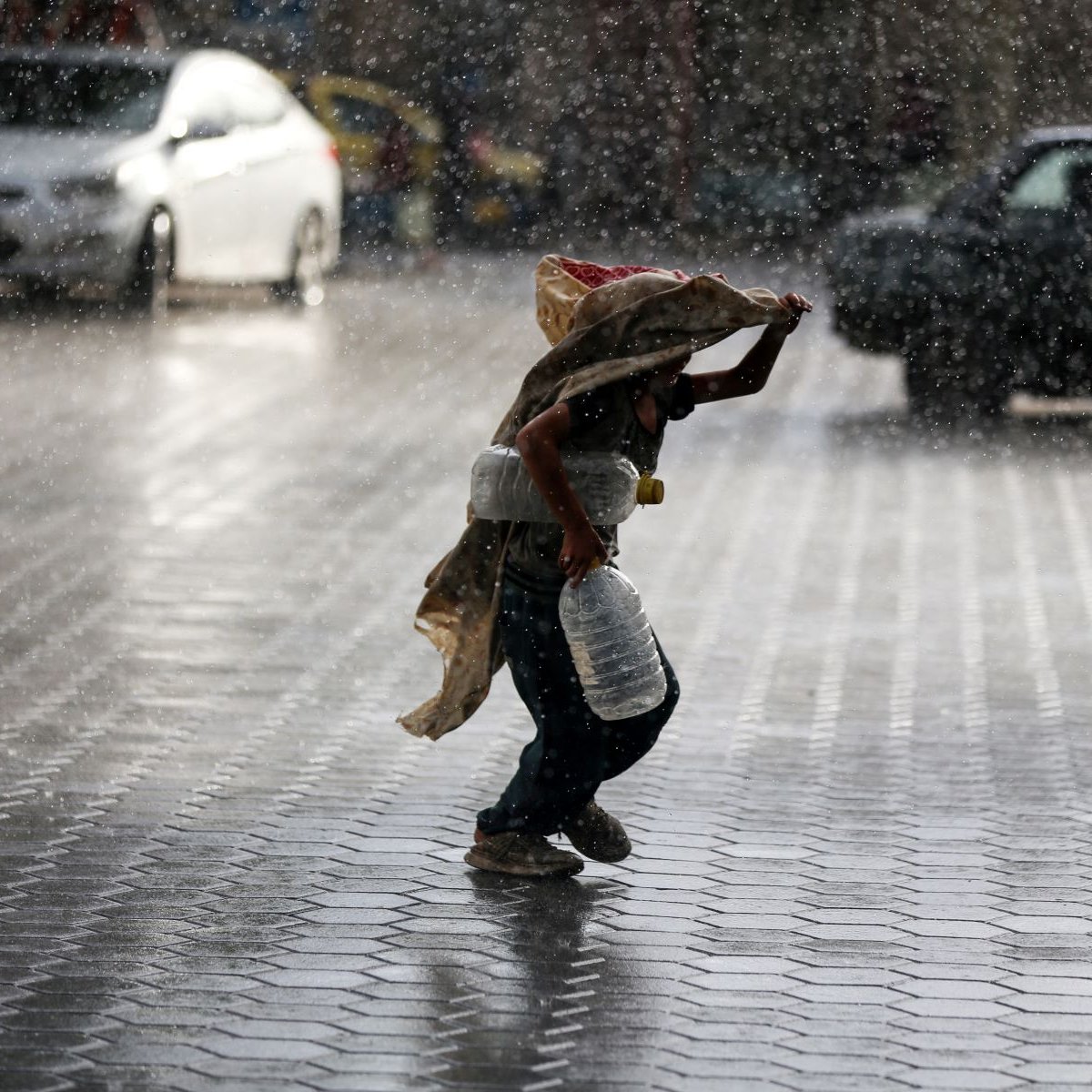 Nell'immagine si vede un bambino che corre sotto la pioggia intensa. Il ragazzo tiene un grande contenitore in una mano e si protegge dalla pioggia con un pezzo di stoffa. Sullo sfondo, ci sono diverse auto e una strada bagnata, mentre le gocce d'acqua creano un'atmosfera drammatica e vivace. La scena trasmette una sensazione di urgenza e resilienza, evidenziando il contrasto tra il bambino e le avversità del tempo.