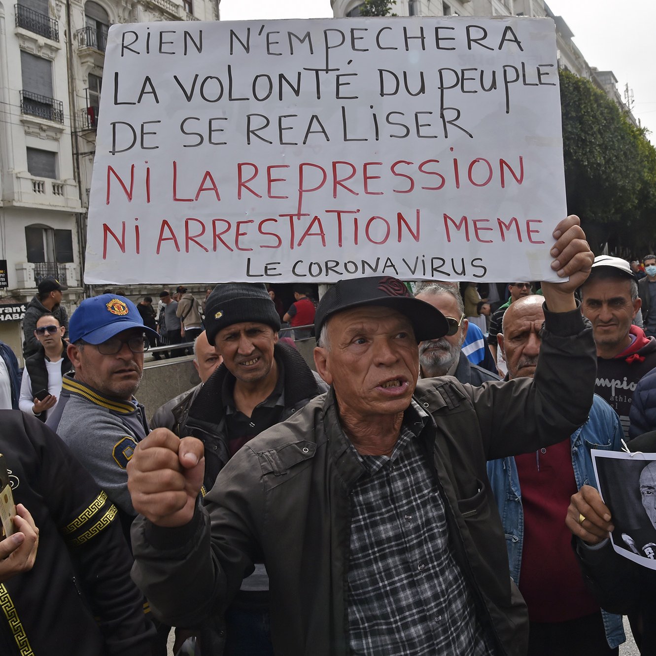 Sur l'image, un groupe de personnes se rassemble dans une rue, probablement lors d'une manifestation. Un homme tient une grande pancarte sur laquelle est écrit : "Rien n'empêchera la volonté du peuple de se réaliser ni la répression ni arrestation même le coronavirus". L'assemblée semble déterminée, et des expressions sérieuses sont visibles sur les visages des manifestants. L'ambiance évoque une lutte pour des droits ou des changements sociaux, sur fond de contestation.