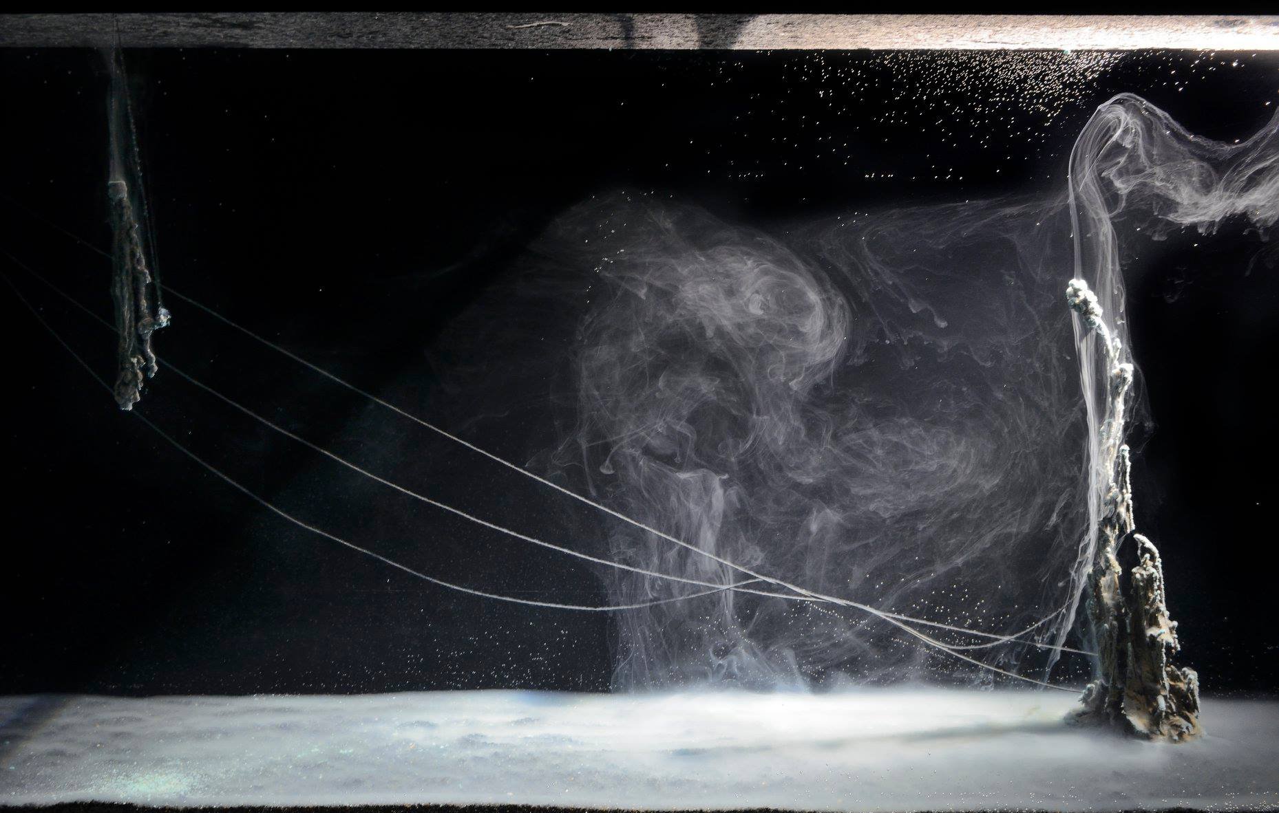 The image showcases a mesmerizing underwater scene featuring swirling currents and bubbles. The foreground has a smooth, almost glass-like surface with a hint of foam, while the background features abstract formations that resemble smoke or flowing water. Thin, delicate tendrils or strings extend across the scene, creating an ethereal atmosphere. The overall color palette is dominated by dark tones, emphasizing the luminous qualities of the water and creating a sense of depth and motion.