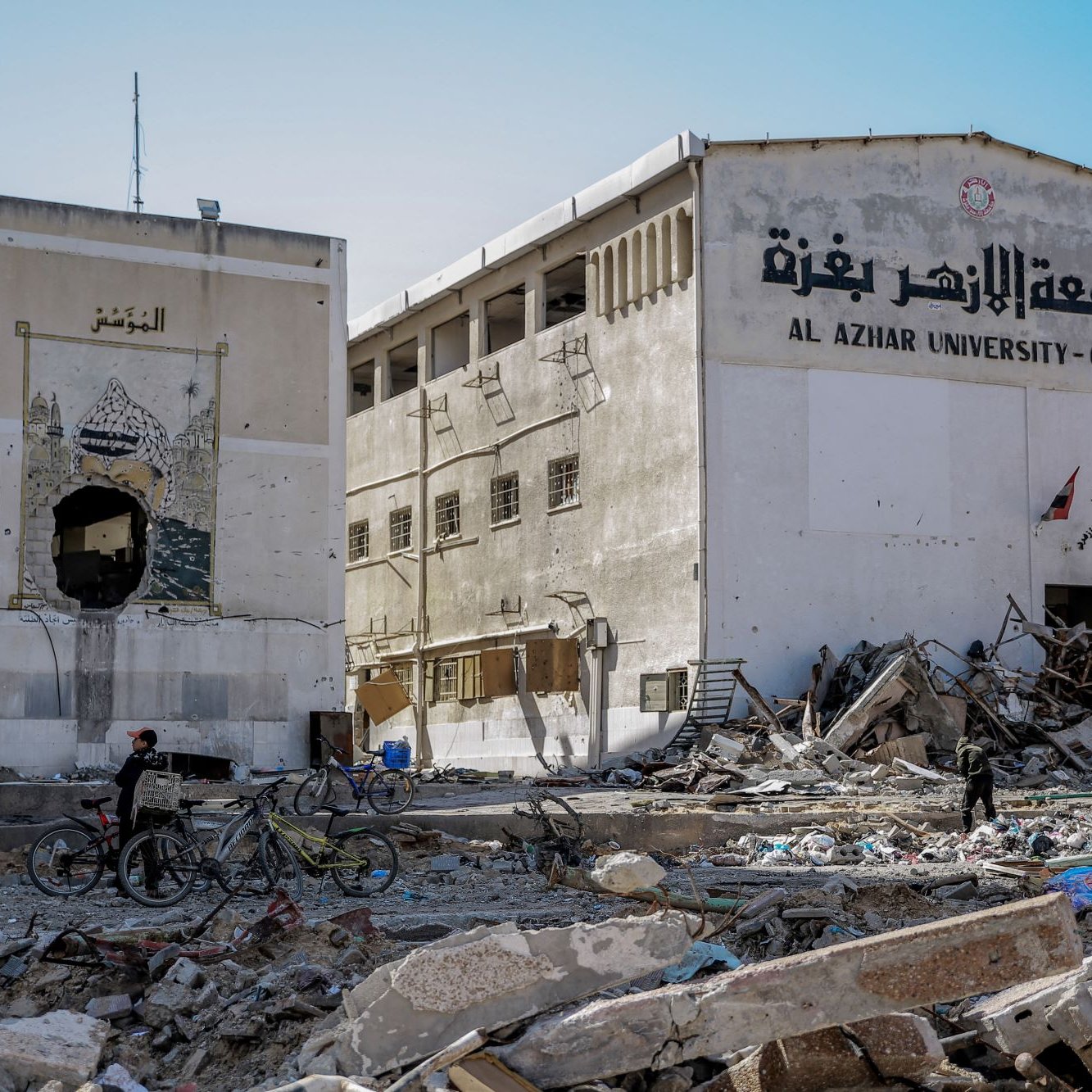 L'immagine mostra un edificio in rovina, probabilmente parte dell'Università Al Azhar di Gaza. La struttura presenta segni di distruzione, con macerie sparse e un'atmosfera di desolazione. A sinistra, si può notare un affresco o un murales deteriorato. Due persone si trovano nel campo visivo, una delle quali è in bicicletta. La scena riflette i danni significativi causati da conflitti e la situazione difficile in quella regione.