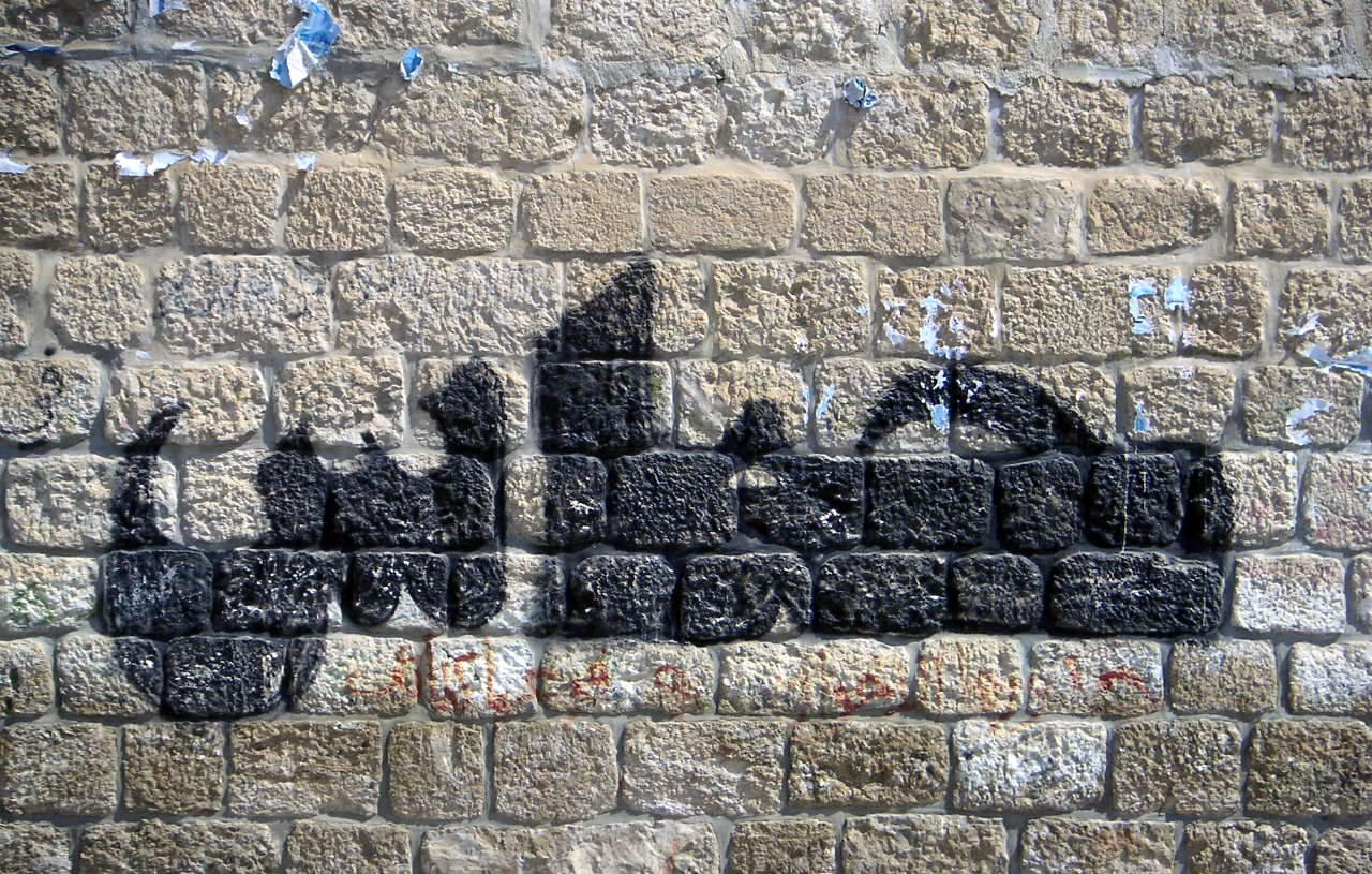 L'image montre un mur de pierres avec un graffiti en forme de silhouette noire. Le mur semble ancien et légèrement abîmé, avec des morceaux de papier déchirés. La silhouette créée par le graffiti évoque des formes humaines, mais sans détails précis. Les pierres du mur sont bien visibles et ajoutent une texture rustique à l'ensemble.