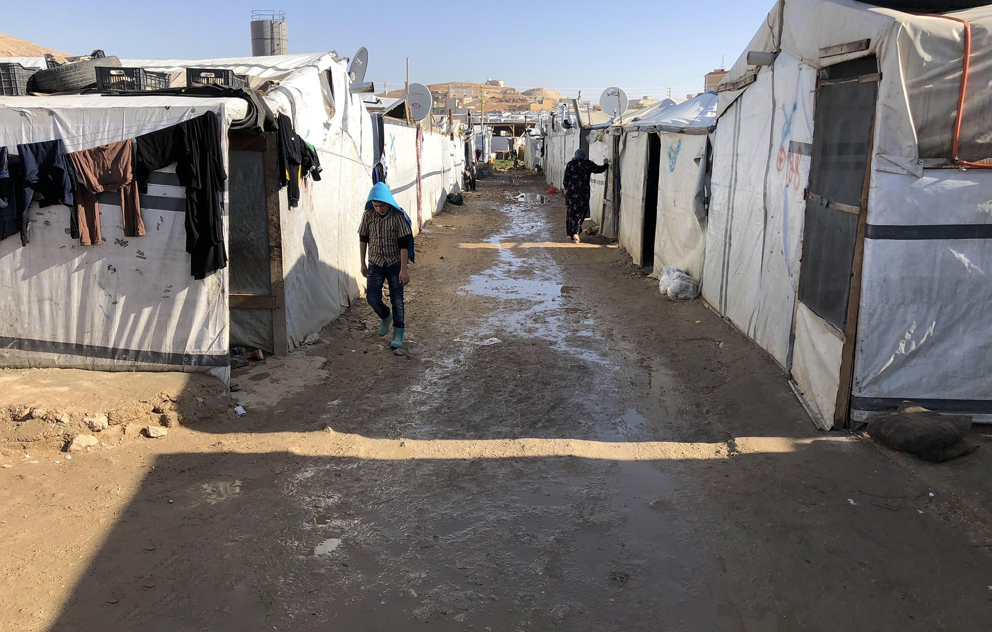 تُظهر الصورة منظرًا لمخيم يتكون من خيام بيضاء متراصة على جانبي ممر ضيق. يبدو أن الأجواء قد كانت ممطرة، حيث تتجمع برك صغيرة من الماء على الأرض. يوجد أشخاص يسيرون في الممر، وبعض الملابس معلقة على الحبال خارج الخيام. يسيطر الشعور بالبساطة والصرامة على البيئة المحيطة، مما يعكس ظروف الحياة في المخيم.