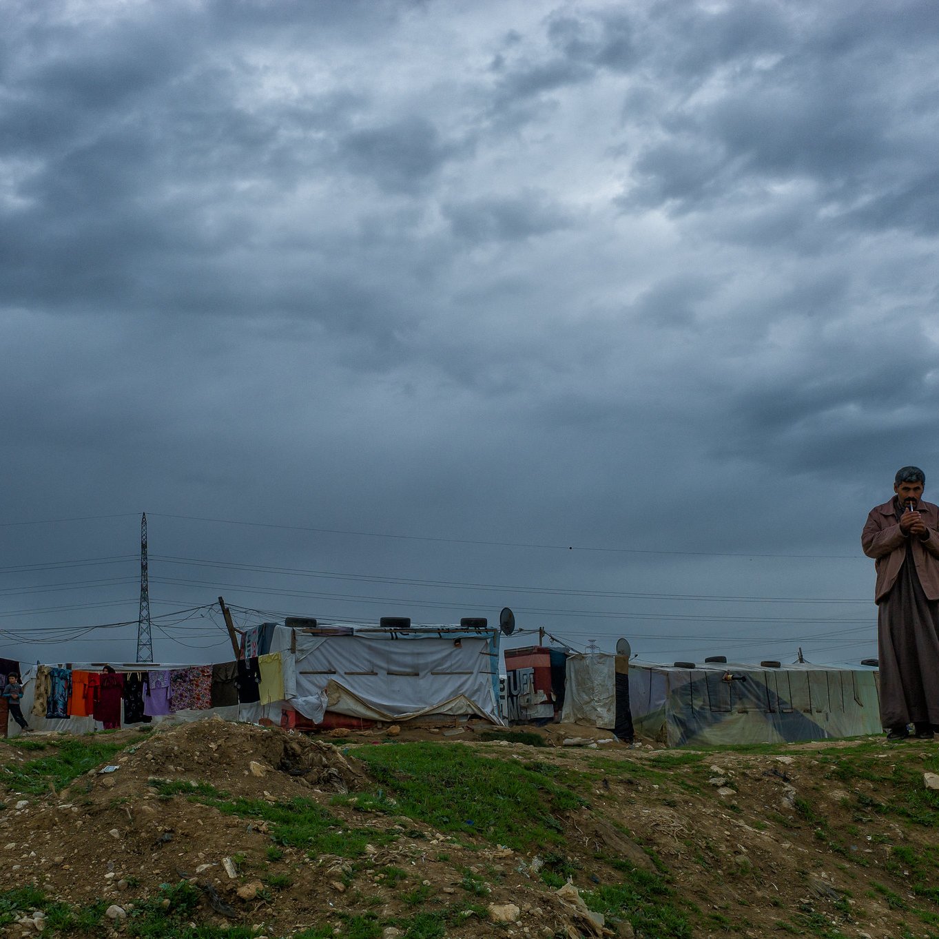 The image depicts a somber landscape under a cloudy sky. In the foreground, a lone figure stands, appearing pensive or contemplative. The background features makeshift structures, likely tents or shanties, indicating a temporary or informal settlement. These structures are adorned with various colors and materials, reflecting a harsh living environment. The overall atmosphere conveys a sense of solitude and resilience against the backdrop of an overcast sky.