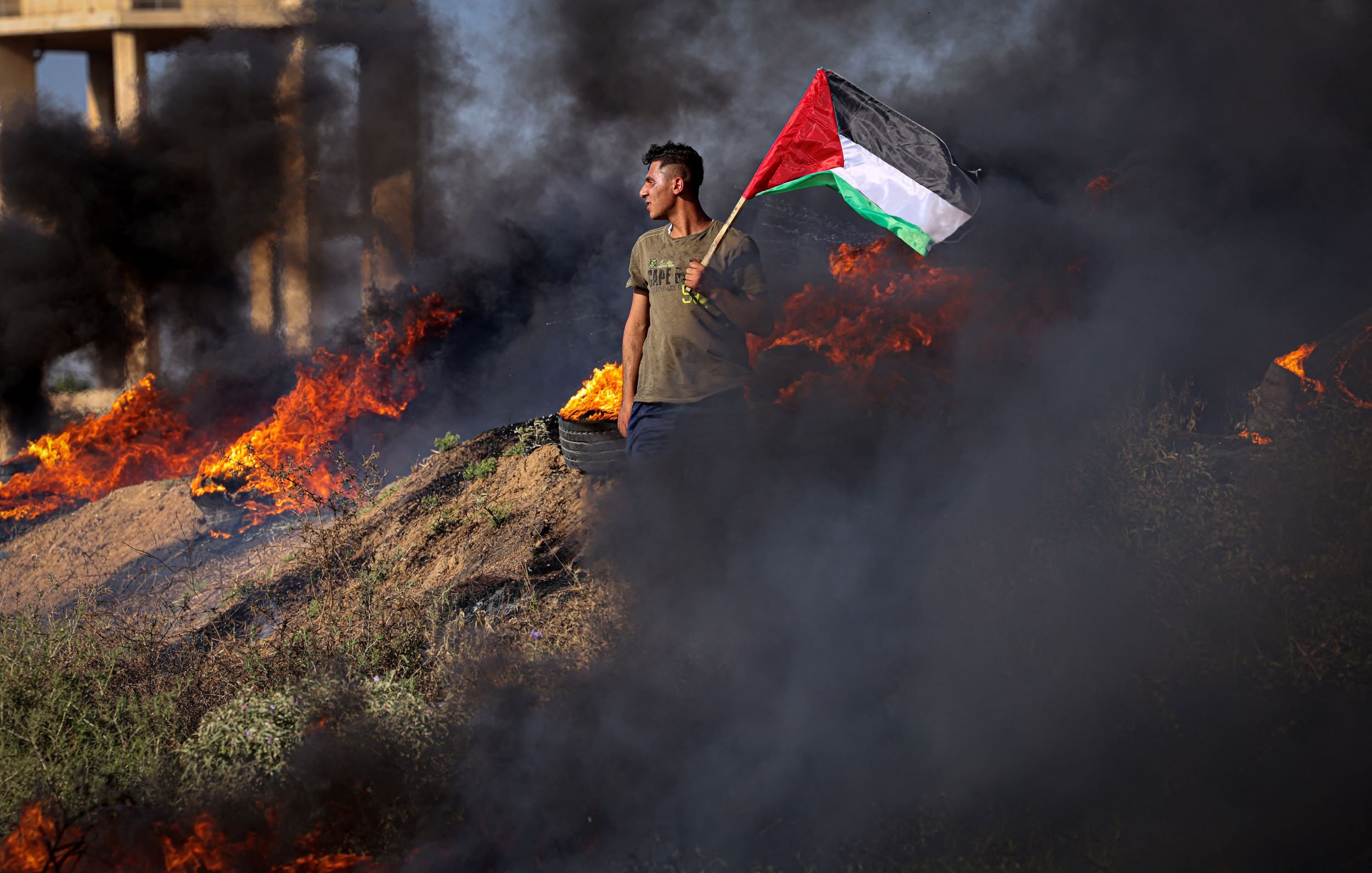 L'image montre une personne debout sur un terrain où des feux brûlent, produisant une épaisse fumée noire. Cette personne tient fièrement un drapeau palestinien, qui est composé de bandes rouge, noire, blanche et verte. Le paysage semble chaotique en raison des flammes entourant le manifestant, créant une atmosphère intense. Les éléments visuels de l'image évoquent des sentiments de militantisme et de détermination.