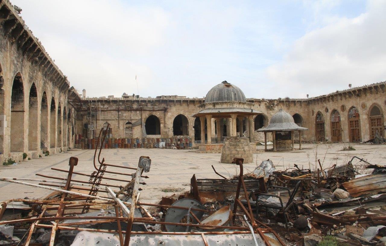 L'image montre un site historique en ruines. On peut voir une cour spacieuse entourée de murs en pierre, avec des arches et des colonnes qui témoignent d'une architecture ancienne. Au centre, il y a deux structures en forme de dômes. Le sol est recouvert de débris métalliques et de morceaux de ferraille, suggérant que le lieu a été touché par des événements destructeurs. Le ciel est nuageux, ajoutant à l'atmosphère de désolation.