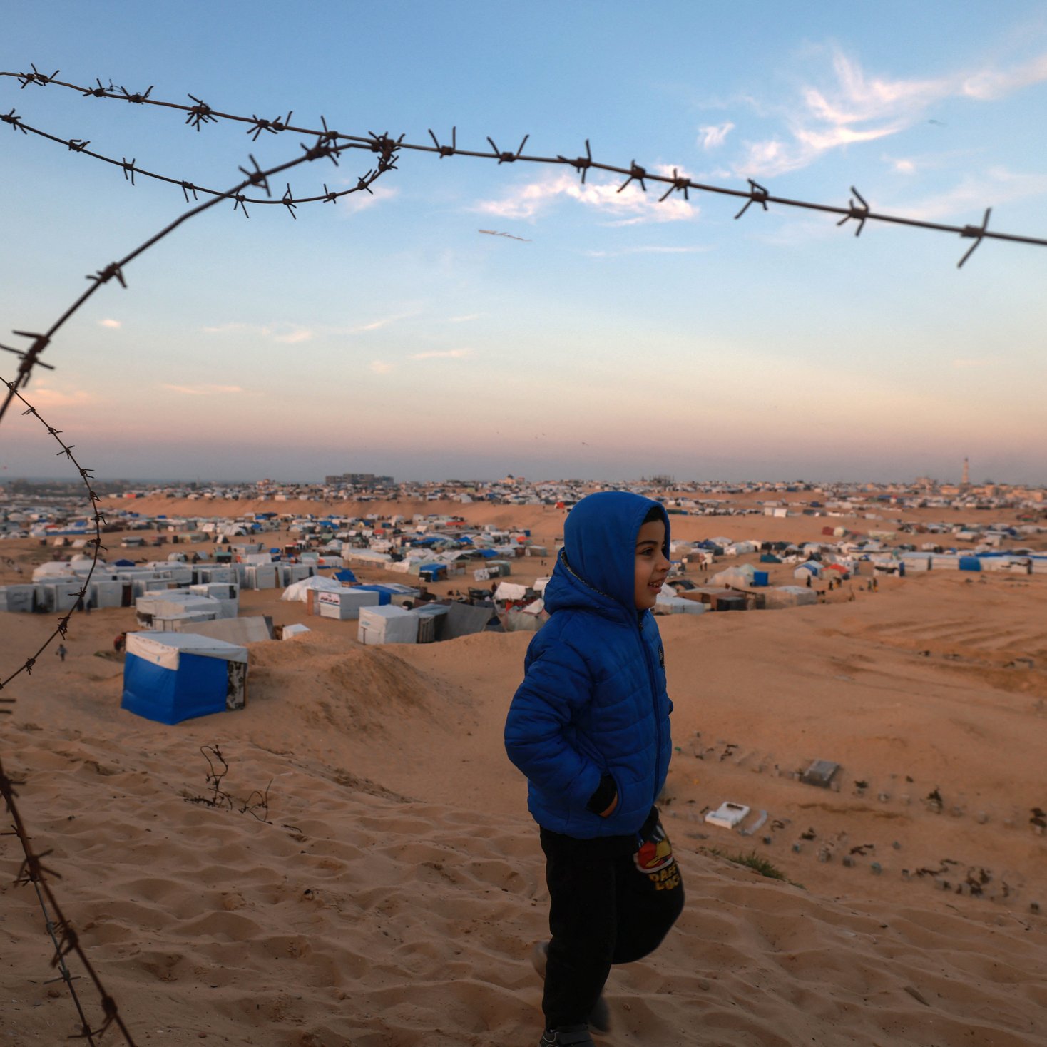 L'image montre un jeune garçon portant une veste bleue qui marche dans un paysage désertique. En arrière-plan, on aperçoit un camp de réfugiés avec de nombreuses tentes et abris disposés sur le sol sablonneux. Un barbelé est visible au premier plan, ajoutant une dimension de sécurité ou de séparation à la scène. Le ciel est partiellement dégagé, avec des teintes de bleu et d'orange, suggérant peut-être la fin de la journée. L'ensemble transmet une atmosphère à la fois de vulnérabilité et d'espoir.