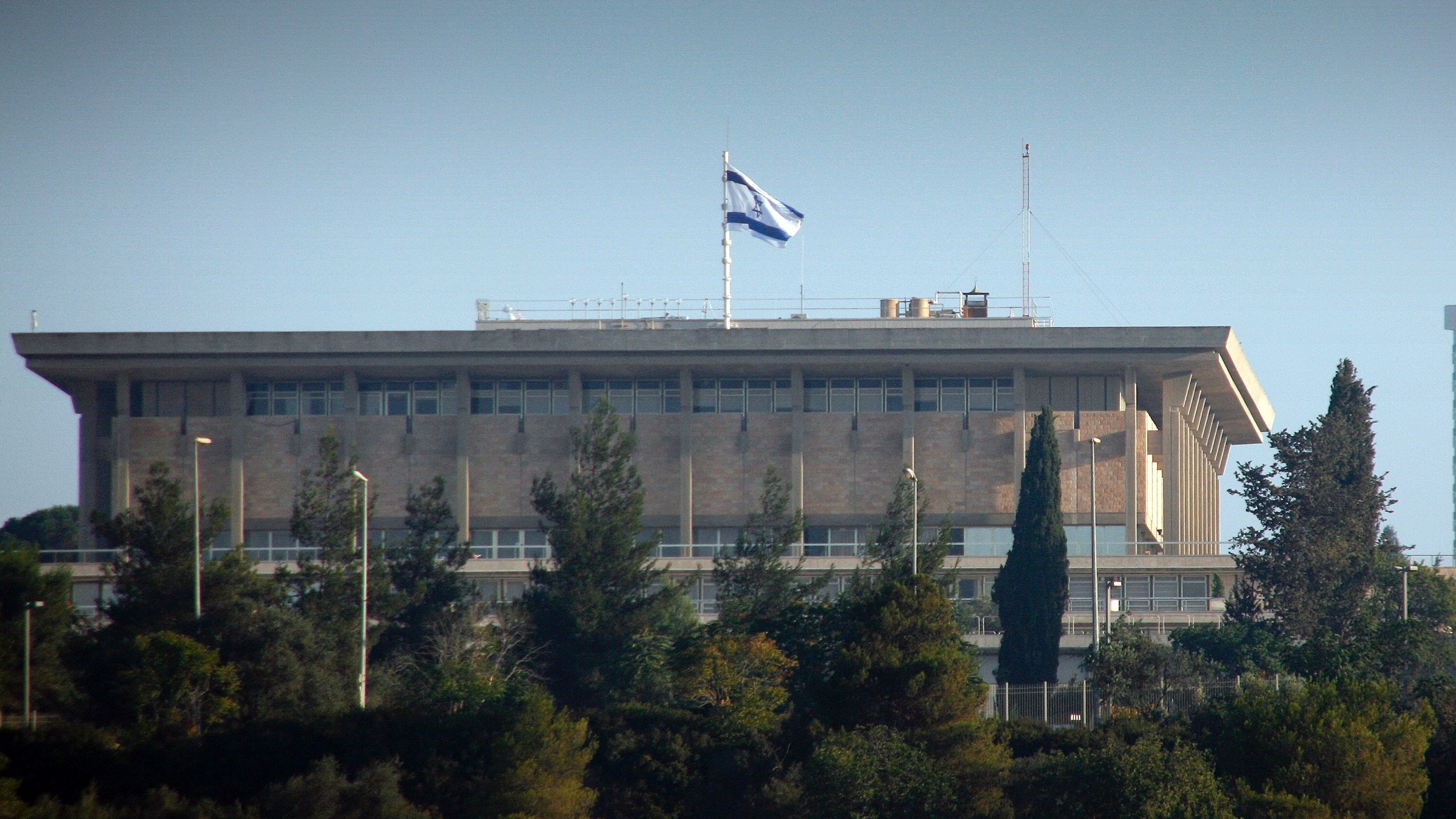 L'image montre un bâtiment imposant, probablement un bâtiment gouvernemental, situé sur une colline. On peut voir le drapeau d'Israël flotter au sommet du bâtiment. Celui-ci est entouré de verdure et d'arbres, indiquant que l'environnement est naturel et paisible. La structure a un style architectural moderne avec de grandes fenêtres et des balcons. L'éclairage suggère que la photo a été prise pendant la journée.