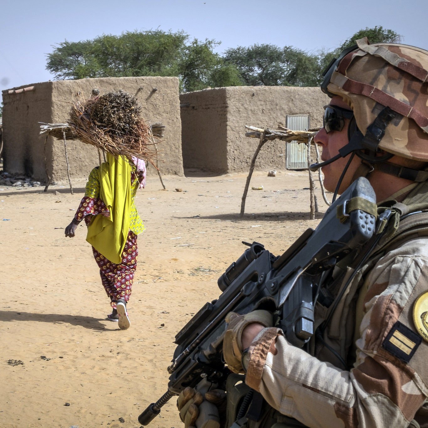 L'image montre un soldat armé en position de surveillance dans un environnement désertique. Il est vêtu d'un uniforme militaire et porte un casque. À l'arrière-plan, on aperçoit une femme portant un fardeau, sans doute des branches, qui s'éloigne pour se rendre à un village constitué de constructions en terre. Le terrain est sablonneux, et quelques arbres épars se trouvent aux alentours. Cette scène évoque un contexte de sécurité dans une région rurale.