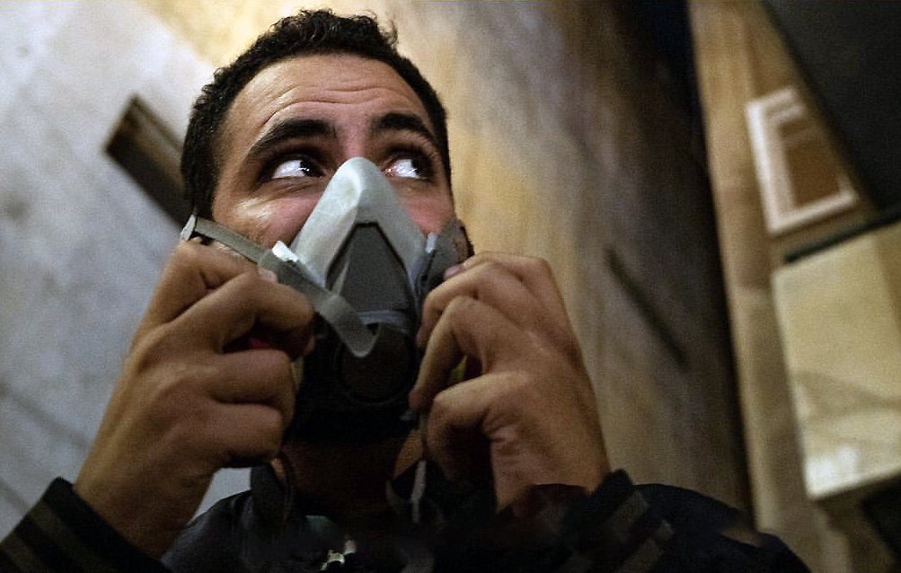 L'image montre un homme qui porte un masque à gaz. Son visage exprime une certaine vigilance, et il semble regarder autour de lui. L'environnement est urbain, avec des murs et des bâtiments en arrière-plan. L'éclairage donne une ambiance sombre et intense à la scène.