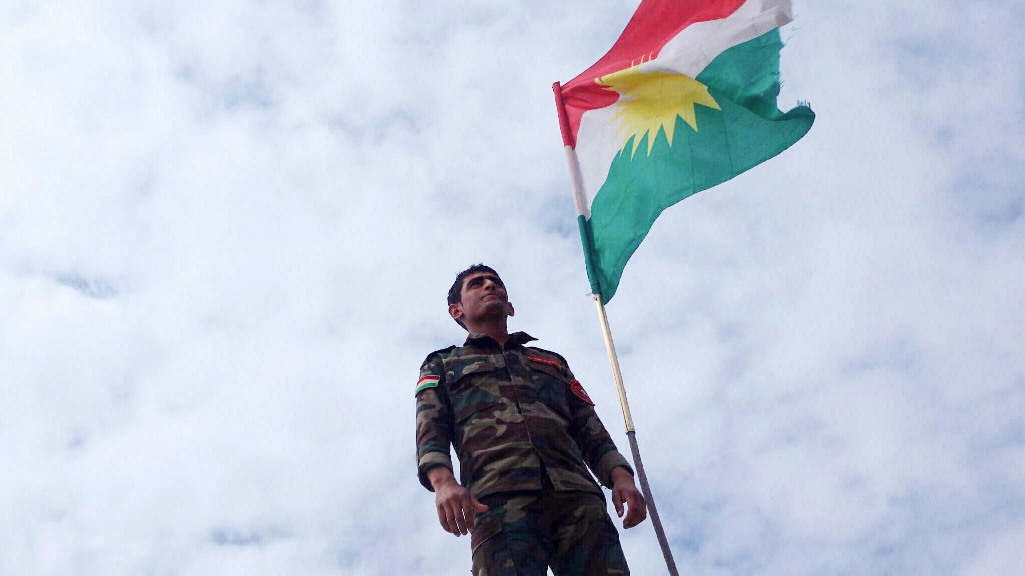L'image montre un soldat portant un uniforme militaire, se tenant debout sous un drapeau kurde. Le drapeau présente les couleurs rouge, vert et blanc avec un soleil rayonnant au centre. Le ciel est nuageux, ajoutant une ambiance dramatique à la scène. Le soldat semble fier et déterminé, symbolisant l'engagement et l'identité kurde.