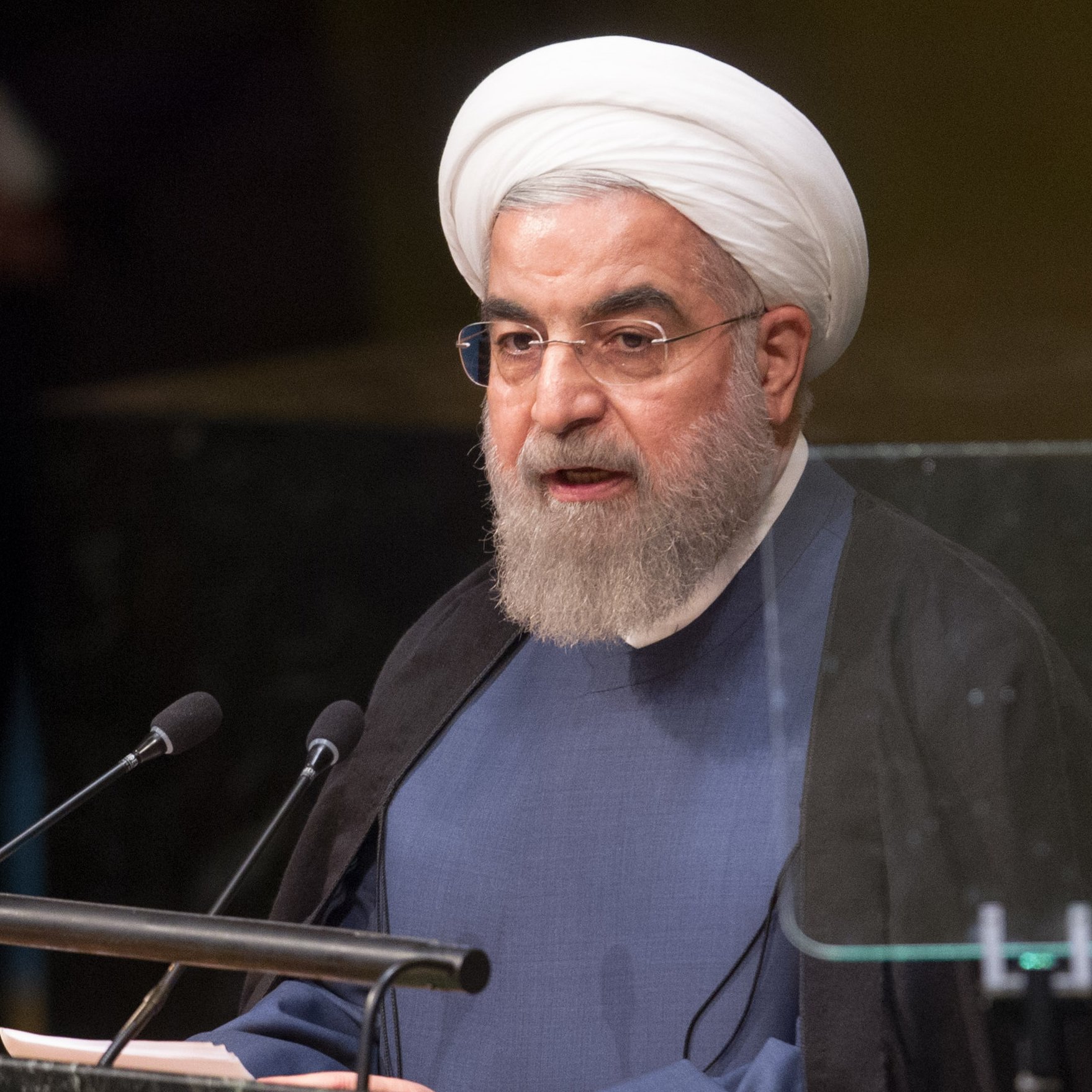 الصورة تظهر شخصًا يتحدث أمام منصة رسمية، ويبدو أنه في موقف رسمي أو مؤتمر. يرتدي هذا الشخص عمامة بيضاء وثوبًا شرعيًا، مما يشير إلى أنه قد يكون شخصية دينية أو سياسية. يتواجد ميكروفون أمامه، ويبدو أنه ينظر إلى الجمهور أو إلى الكاميرات. الخلفية غامضة قليلاً، مما يضيف إلى الطابع الرسمي للمشهد.