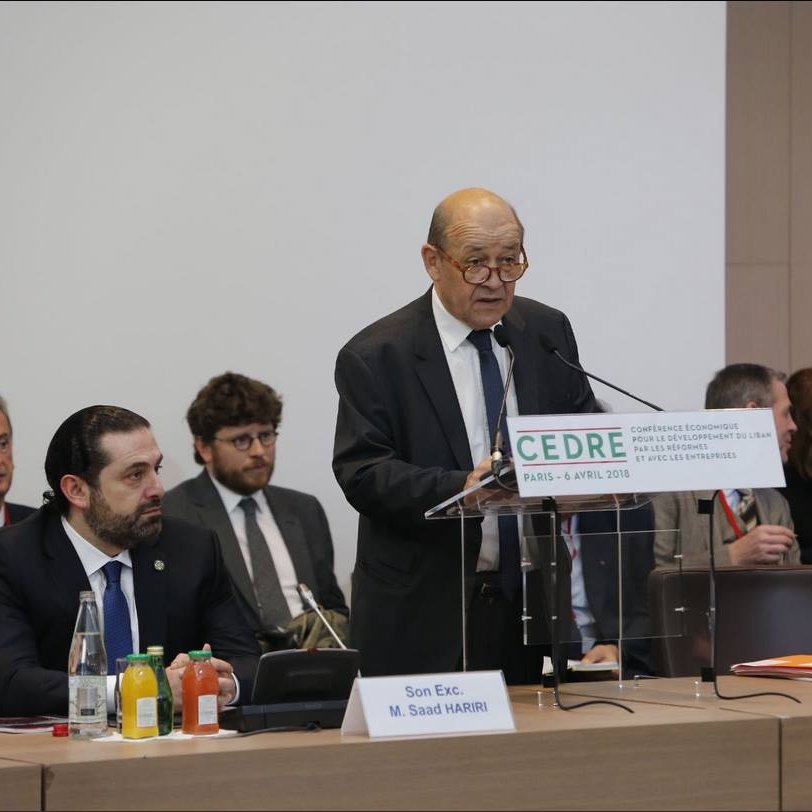 The image depicts a formal conference setting. A middle-aged man, presumably a speaker, is standing at a podium with a sign that reads "CEDRE" and indicates the location and date of the event. He appears to be addressing an audience that includes several men seated behind him, some of whom are engaged in listening or taking notes. The environment has a professional tone, with tables set up and various drinks and papers visible. The atmosphere suggests a significant discussion or presentation, likely focused on economic or policy-related matters.