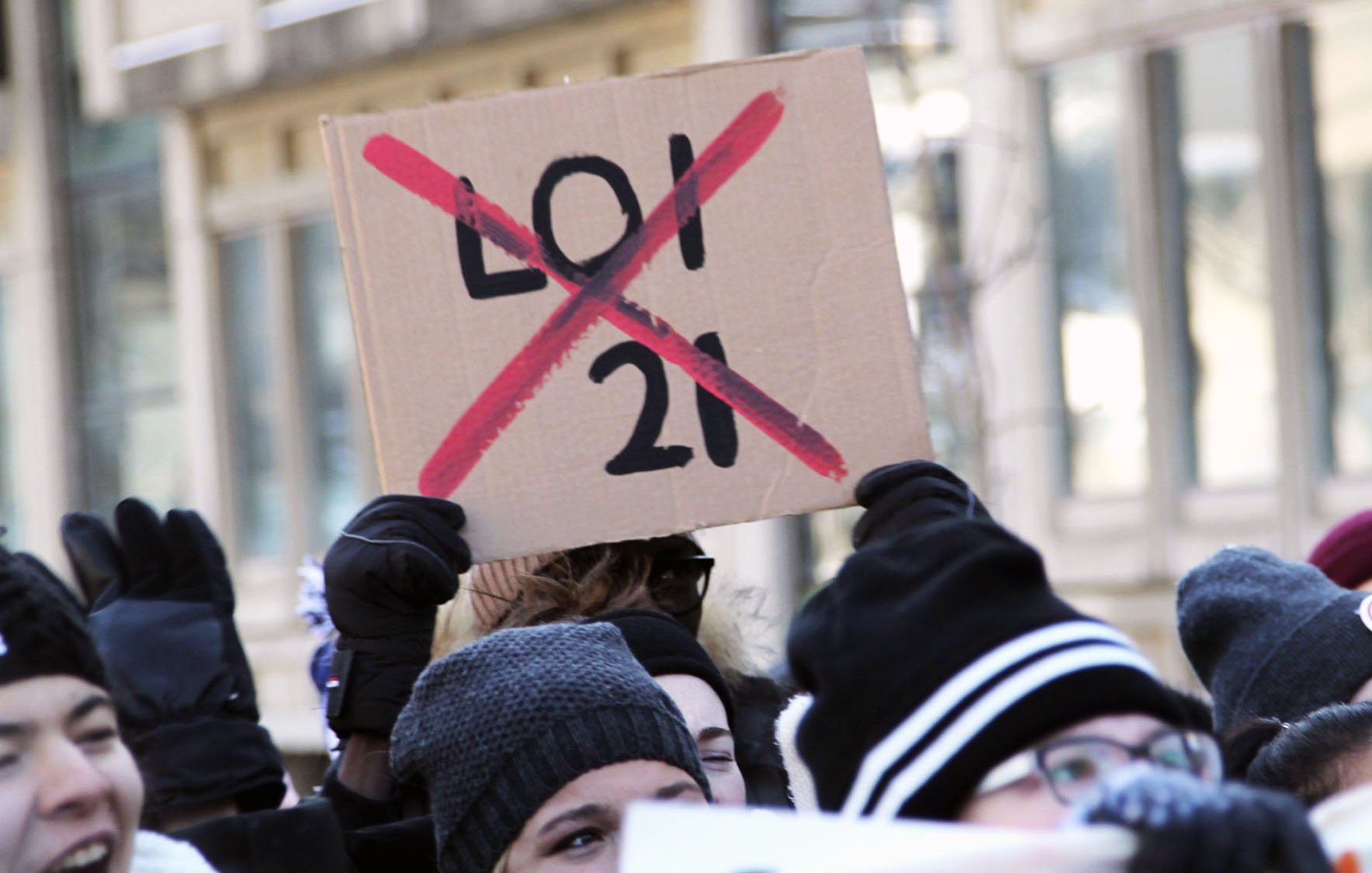 La imagen muestra a un grupo de personas participando en una manifestación. Algunas de ellas sostienen carteles, y uno de los más destacados es un cartel de cartón que dice "X" sobre el número "21", lo que indica oposición a esa ley o número específico. La multitud parece estar animada y comprometida con la causa que defienden. Las personas están abrigadas, usando gorros y guantes, lo que sugiere que hace frío.