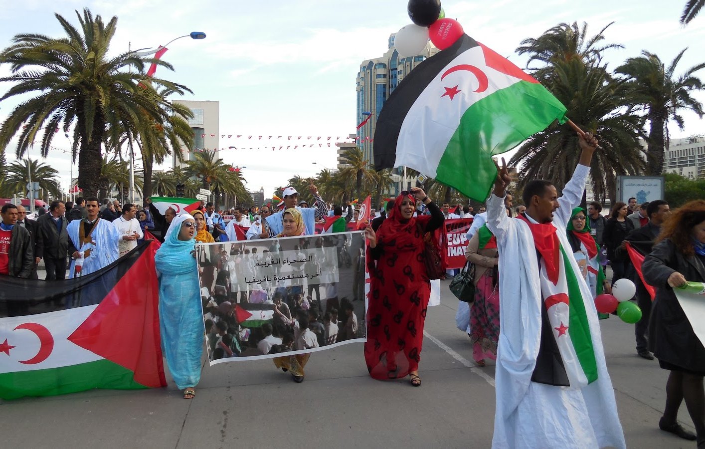 The image depicts a lively demonstration, likely in support of a cause related to Western Sahara. People are marching along a street, holding banners and flags that feature the colors red, green, and black, which are associated with the Sahrawi nationalist movement. The crowd includes both men and women, some wearing traditional clothing. In the background, palm trees and buildings suggest a warm climate, and festive decorations, like balloons, add to the atmosphere of the event. The banner appears to convey a message related to their cause.