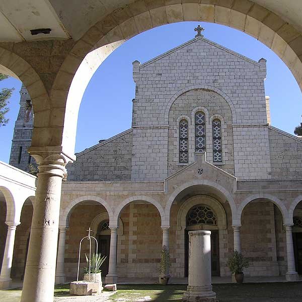 L'image montre un cloître avec des arches stylisées entourant un jardin. Au centre, on peut voir une structure architecturale en pierre, probablement une église ou un monastère, avec un toit en pente et des fenêtres ornées. L'ambiance est paisible, avec une lumière naturelle qui illumine le décor, créant un contraste entre l'ombre des arches et la clarté extérieure. Des éléments de verdure, comme des plantes, ajoutent une touche de vie à l'environnement.