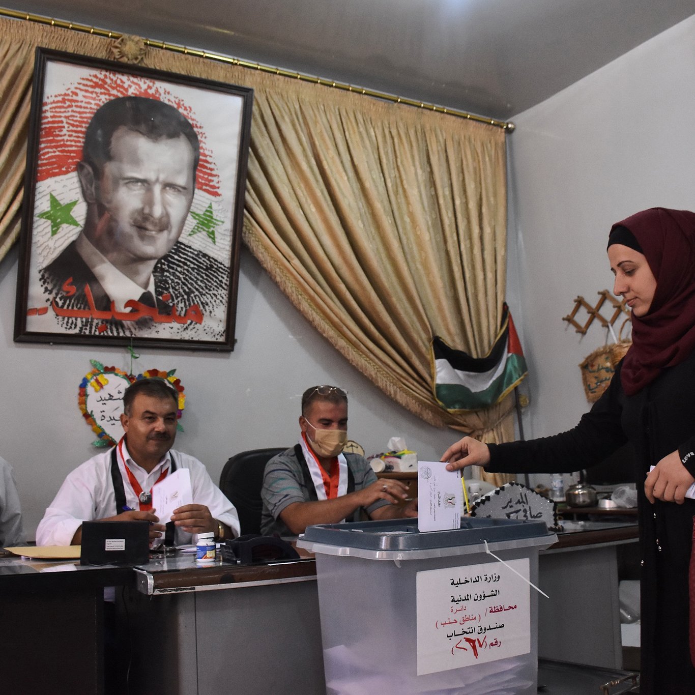 تظهر الصورة امرأة ترتدي عباءة وحجابًا تضع ورقة اقتراع في صندوق الانتخاب. خلفها، يوجد لوحة كبيرة تحمل صورة شخصية معروفة. يجلس ثلاثة رجال خلف طاولة، حيث يبدو عليهم الاستعداد لمراقبة العملية الانتخابية. المكان يبدو رسميًا، مع ستائر وخلفية تشير إلى الطابع المحلي.