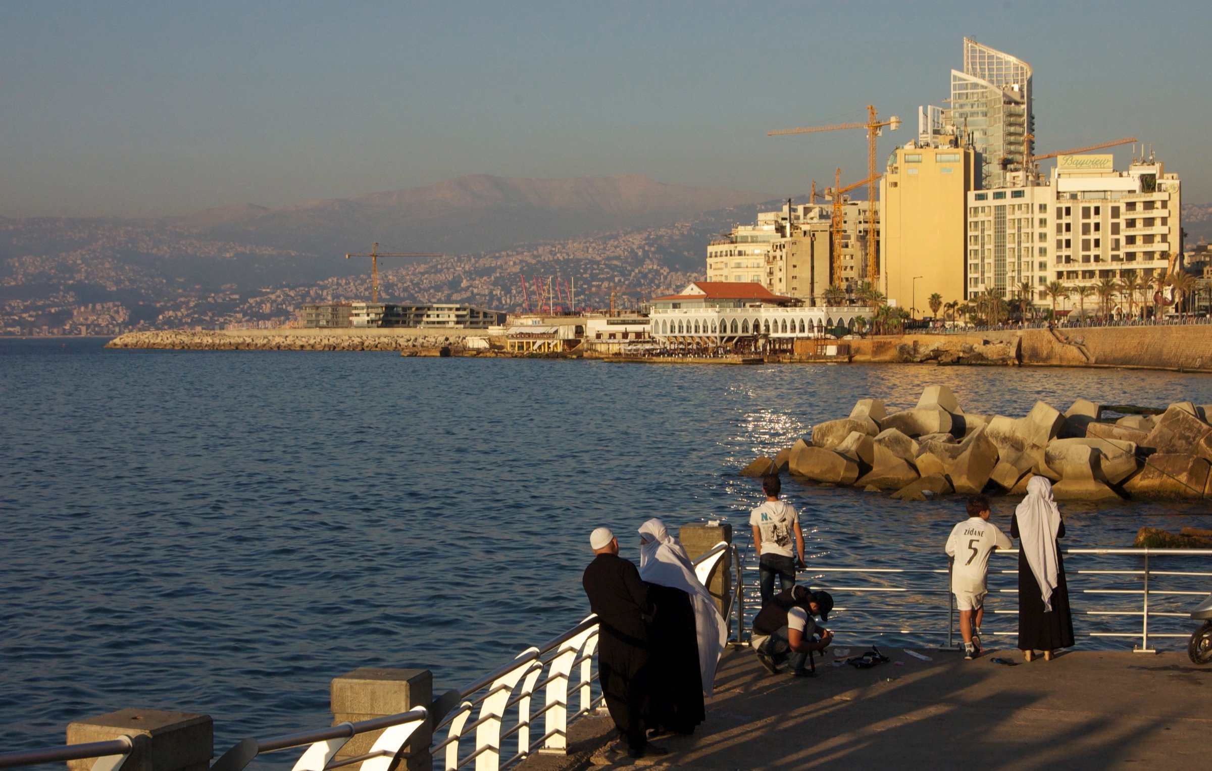 L'image montre un paysage côtier au bord de la mer. On peut voir une promenade avec des personnes qui se tiennent près de l'eau, probablement en train de profiter du coucher de soleil. Au fond, il y a des bâtiments modernes et quelques grues de construction, suggérant un développement urbain. La mer est calme et reflète la lumière dorée du soleil. La scène dégage une atmosphère paisible et agréable.