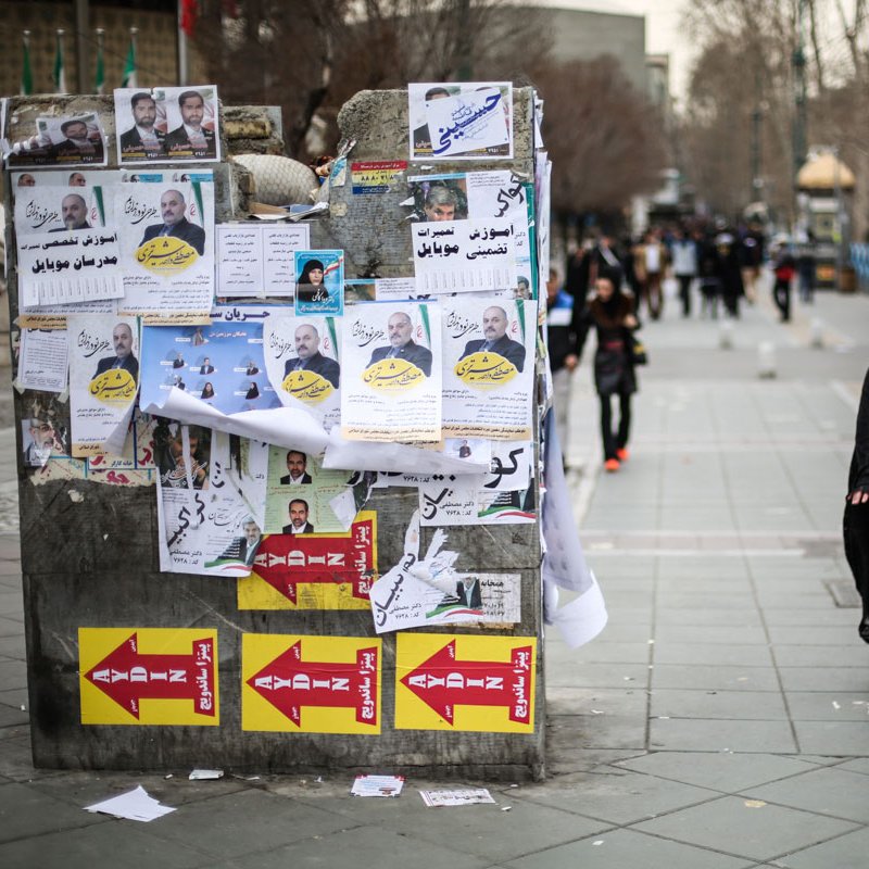 L'image montre une rue animée où une femme portant un hijab noir marche sur le trottoir. À côté d'elle, un mur est recouvert d'affiches et de banderoles, certaines de couleur vive, d'autres jaunies et déchirées. Les affiches semblent inclure des images de personnes et des slogans en écriture arabe. En arrière-plan, on peut distinguer des bus et des passants, créant une atmosphère urbaine. Le sol est en pavé, et les arbres dépouillés suggèrent une saison froide.