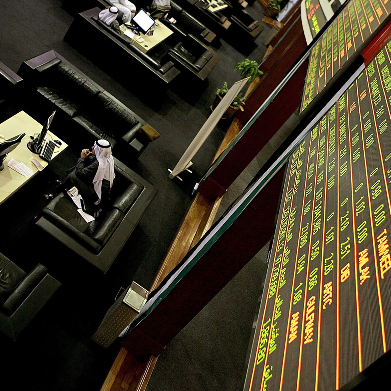 L'image montre un environnement de bourse avec des tables et des sièges confortables. À l'arrière-plan, un grand tableau électronique affiche des informations financières, comme des chiffres et des noms de sociétés. On peut voir quelques personnes, dont un homme en tenue traditionnelle, probablement en train de suivre les fluctuations du marché. L'ambiance semble professionnelle et concentrée, caractéristique d'une salle de négociation.