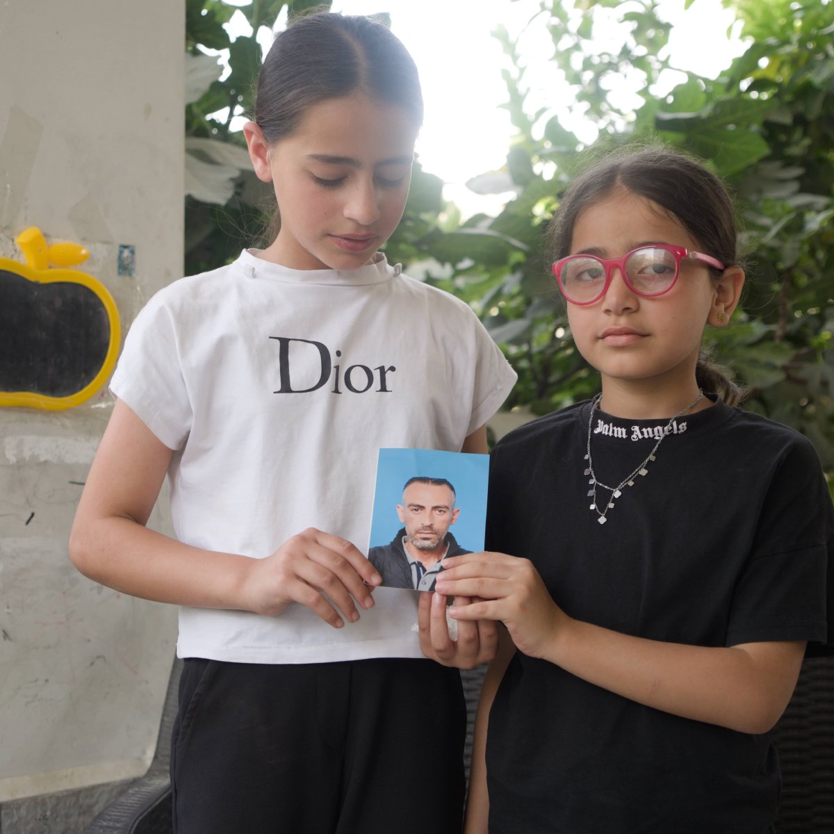 Sur l'image, on voit deux jeunes filles debout l'une à côté de l'autre. La fille de gauche porte un t-shirt blanc avec le logo "Dior" et tient en main une photo d'un homme. La fille de droite est vêtue d'un t-shirt noir avec des lunettes roses. Elles sont entourées par un arrière-plan verdoyant, avec des feuilles visibles autour d'elles.