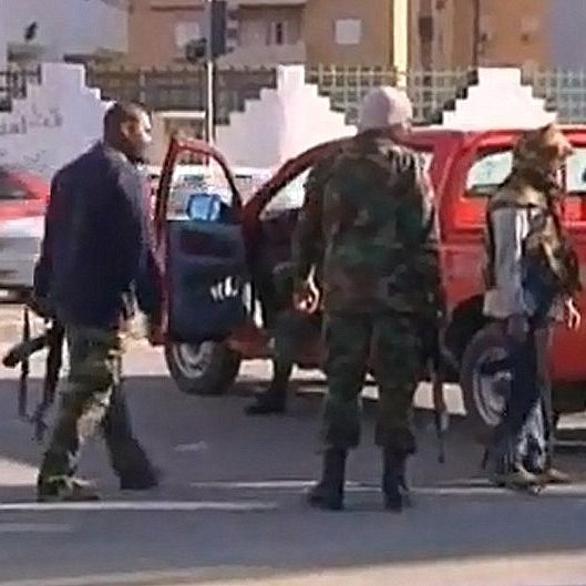 L'image montre une scène de chaos impliquant des personnes en uniforme et armées. Quelques individus se déplacent près de véhicules, tandis qu'un homme semble gesticuler au loin. L'environnement urbain est visible, avec des bâtiments en arrière-plan et une circulation routière. L'atmosphère semble tendue, suggérant un contexte de conflit ou d'incident de sécurité.