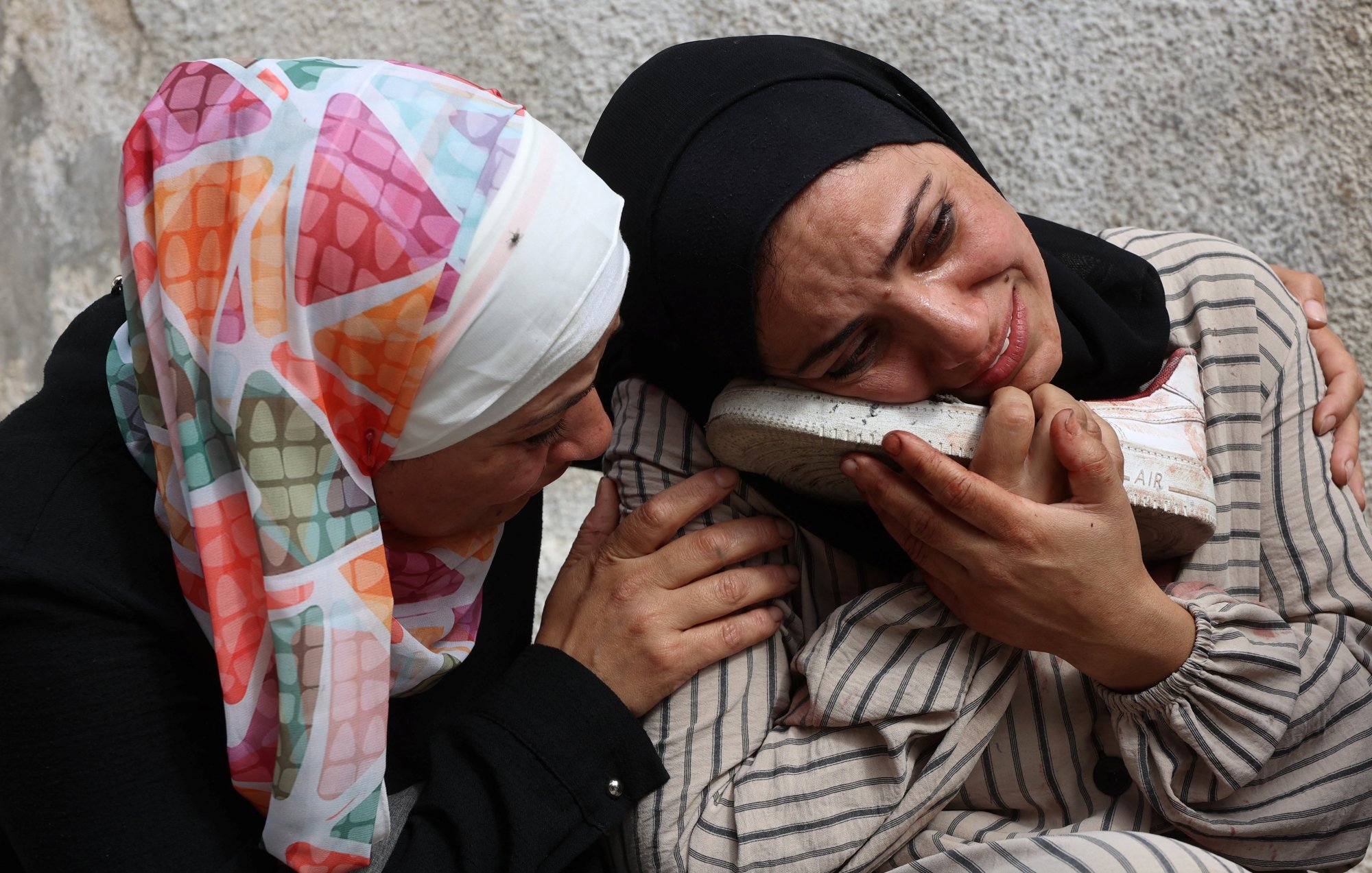 Two women in headscarves are embracing, expressing deep sorrow, with one holding a shoe.
