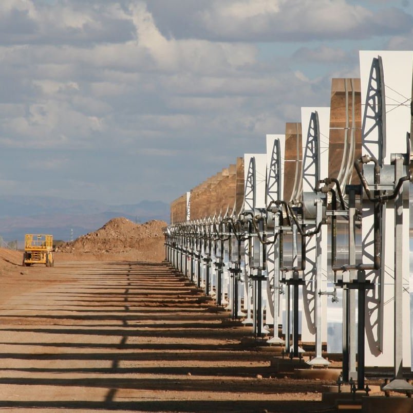 تظهر الصورة مجموعة من الألواح الشمسية المرتبطة بهياكل معدنية، تقع في منطقة صحراوية أو شبه صحراوية. الألواح مرتبة بشكل منتظم وموجهة نحو السماء، مما يشير إلى أنه يجري استغلال الطاقة الشمسية. في الخلفية، يمكن رؤية تلال ترابية وبعض السحب في السماء، مما يعطي انطباعًا عن مساحة واسعة ومفتوحة. تُظهر الصورة أيضًا طريقًا يسير فيه مركبة كبيرة تشير إلى وجود نشاط صناعي أو زراعي في المنطقة.