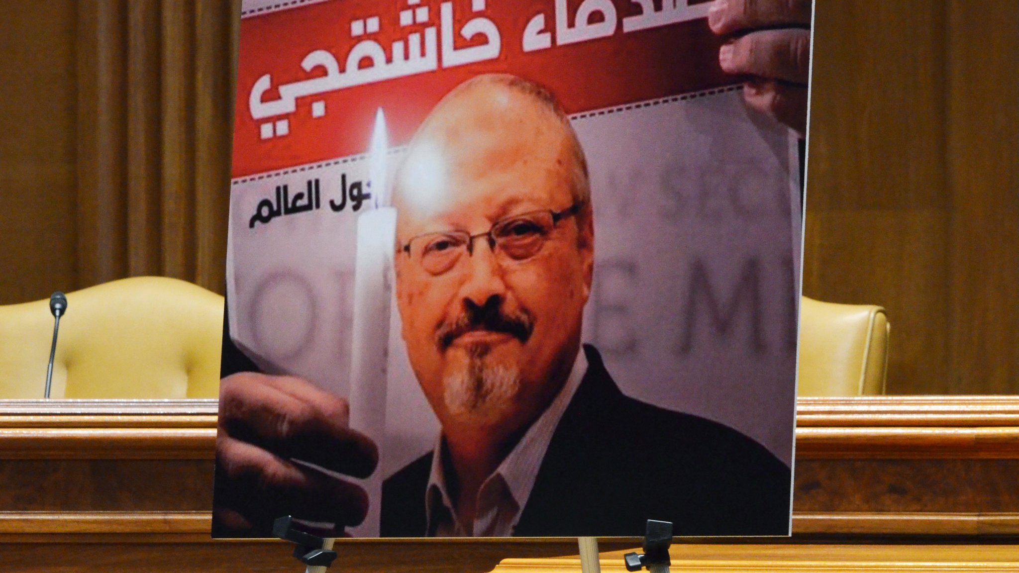 The image features a large poster of a man holding a candle, set against a background with text in Arabic. The poster is likely used in a context related to remembrance or tribute. The setting appears formal, possibly in a legislative or governmental chamber, indicated by the presence of chairs and the overall decor. The image emphasizes themes of justice and memory.