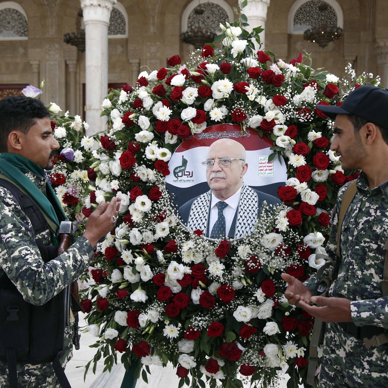 دو نظامی در کنار تاج گل با گل‌های قرمز و سفید ایستاده‌اند. تصویری از یک مرد وسط تاج قرار دارد.