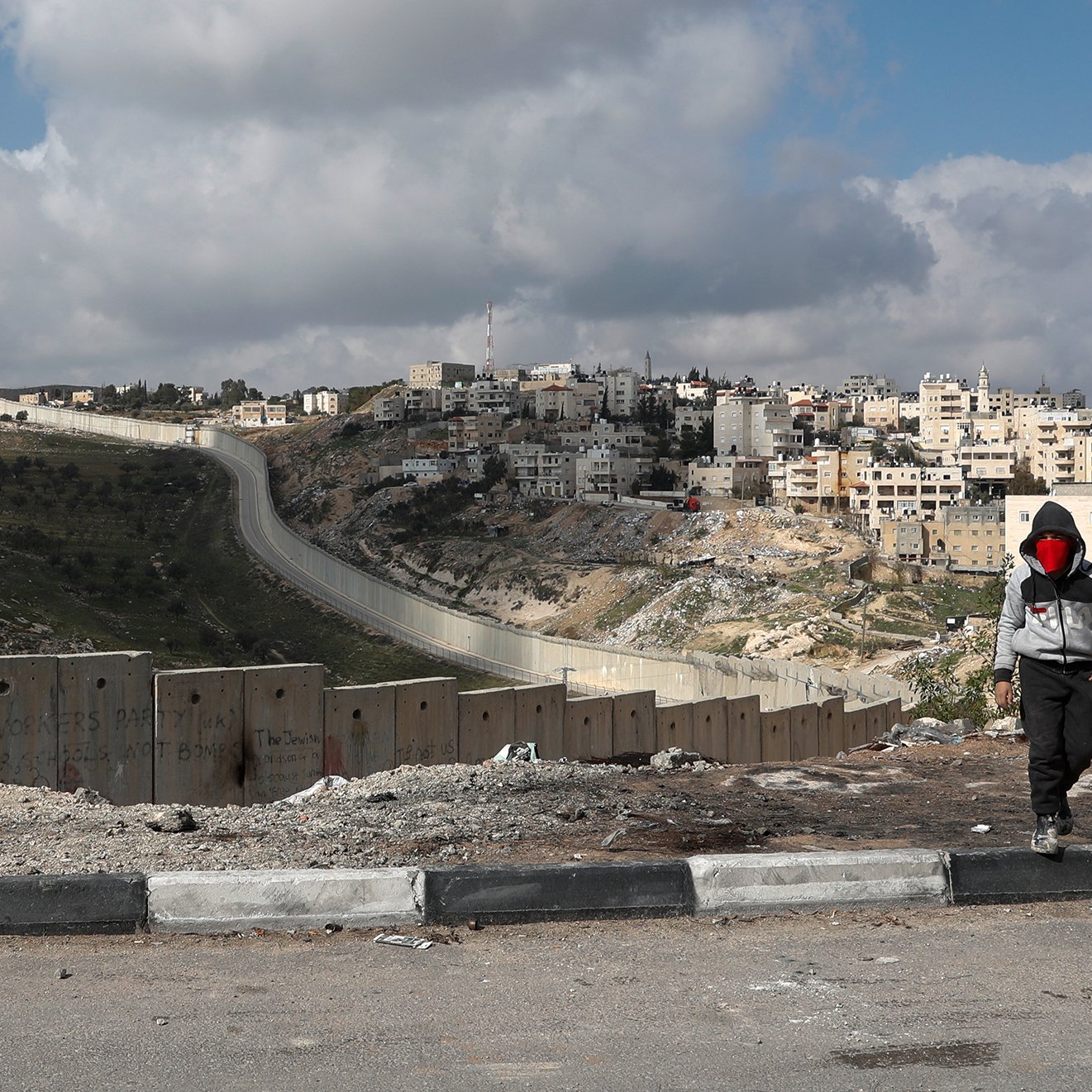 L'image montre un enfant marchant le long d'une route, avec des blocs de béton formant une barrière sur le côté. En arrière-plan, on peut voir une colline surmontée de bâtiments, indiquant une zone urbaine. Le ciel est partiellement nuageux, et le paysage semble aride et rocailleux. L'enfant porte une capuche et un masque, ajoutant une dimension de mystère à la scène. La barrière suggère des thèmes de division et de conflit.