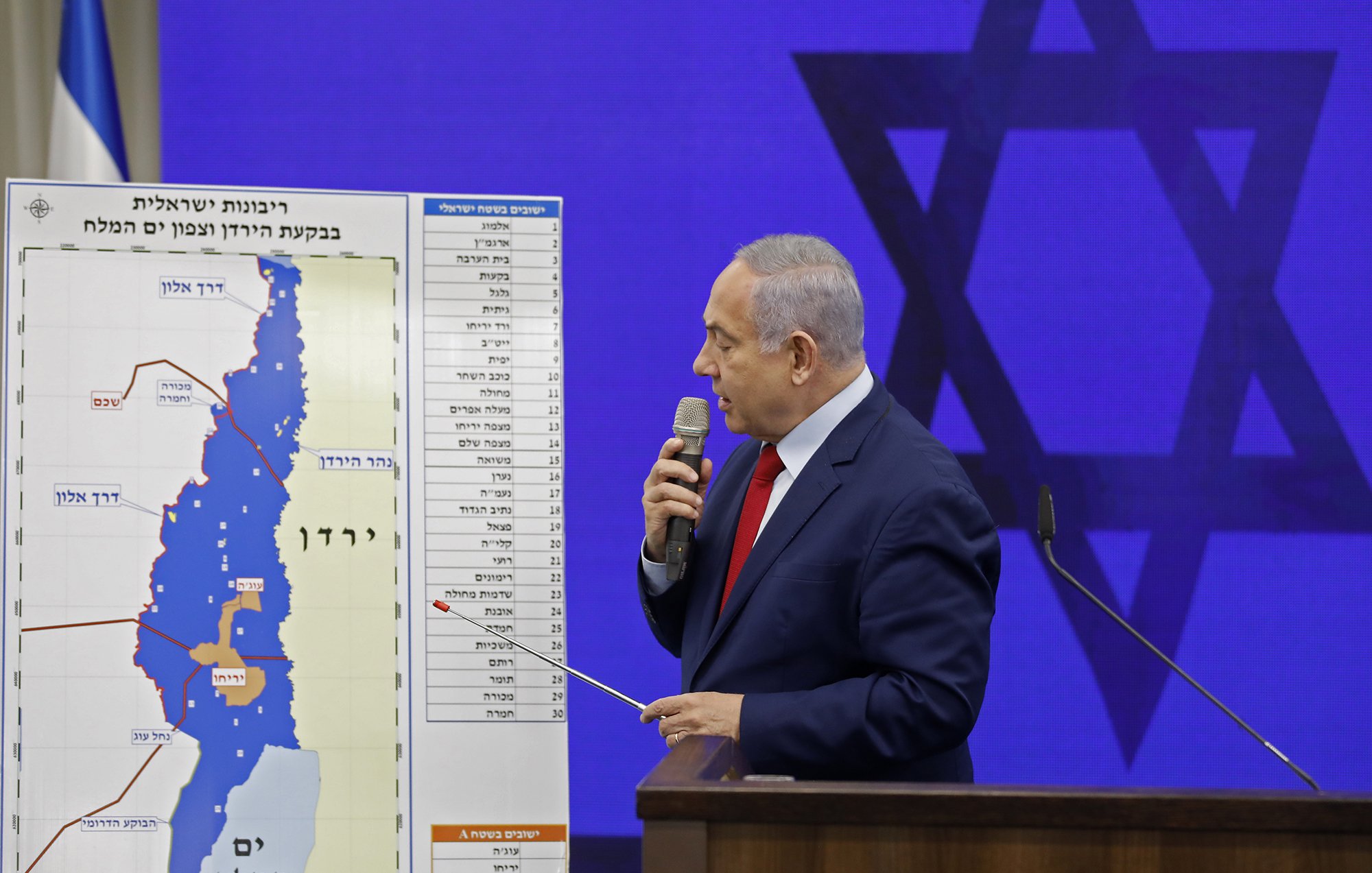 در این تصویر، یک مرد در حال ارائه یک نقشه است. او با یک میلیا بیل کوچک در دست، به نقطه‌ای روی نقشه اشاره می‌کند. نقشه در پس‌زمینه شامل خطوط و علامات جغرافیایی است. پشت او یک علامت بزرگ ستاره داوود دیده می‌شود و فضای سالن به‌نظر می‌رسد رسمی و مرتبط با یک رویداد یا کنفرانس باشد. لباس مرد رسمی به نظر می‌رسد و او به‌وضوح در حال بیان نکات کلیدی است.