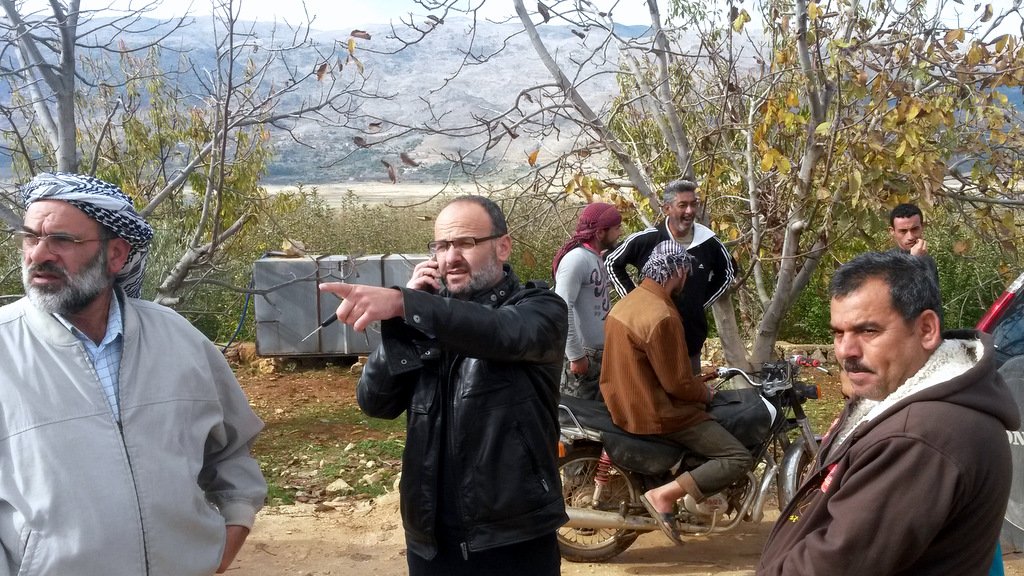 L'image montre un groupe d'hommes dans un environnement rural, probablement en plein air avec des arbres autour. Un homme, habillé avec une veste noire, est au centre, en train de parler au téléphone et de pointer du doigt dans une direction. D'autres hommes sont dispersés autour de lui, certains se tenant debout, d'autres assis sur une moto. L'arrière-plan montre des collines ou des montagnes, suggérant une zone naturelle. L'atmosphère semble détendue mais attentive, comme si les hommes discutaient d'un sujet important.