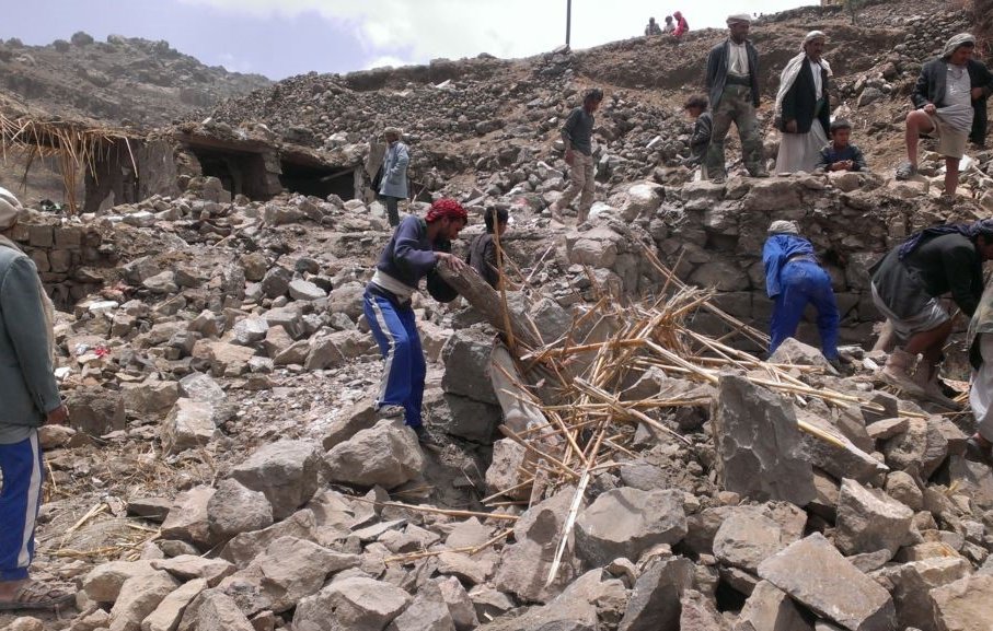 L'image montre un groupe de personnes travaillant dans les décombres d'un bâtiment effondré. Elles semblent s'affairer à dégager des pierres et des débris, avec des outils improvisés. L'environnement paraît montagneux et accidenté, avec des restes de structures anciennes en arrière-plan. L'atmosphère est celle de l'effort collectif dans un contexte de reconstruction ou d'aide humanitaire.