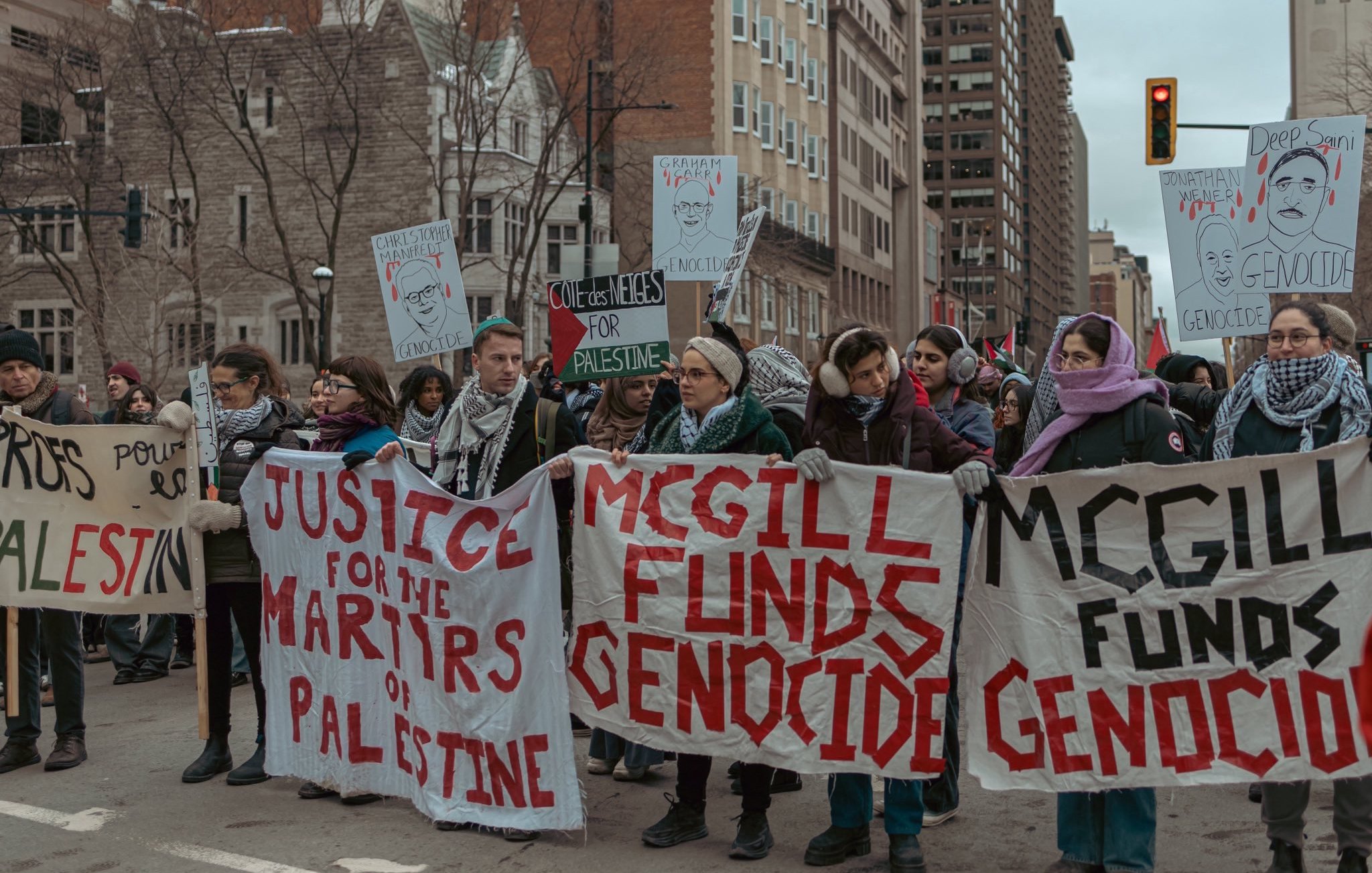 L'image montre une manifestation dans une ville urbaine. Un groupe de personnes est rassemblé, tenant des pancartes avec des messages en faveur de la Palestine et s'opposant au financement de ce qu'ils appellent un génocide. Certains manifestants portent des écharpes qui semblent indiquer leur solidarité avec la cause palestinienne. L'atmosphère est engagée et déterminée, avec des passants et des bâtiments en arrière-plan.