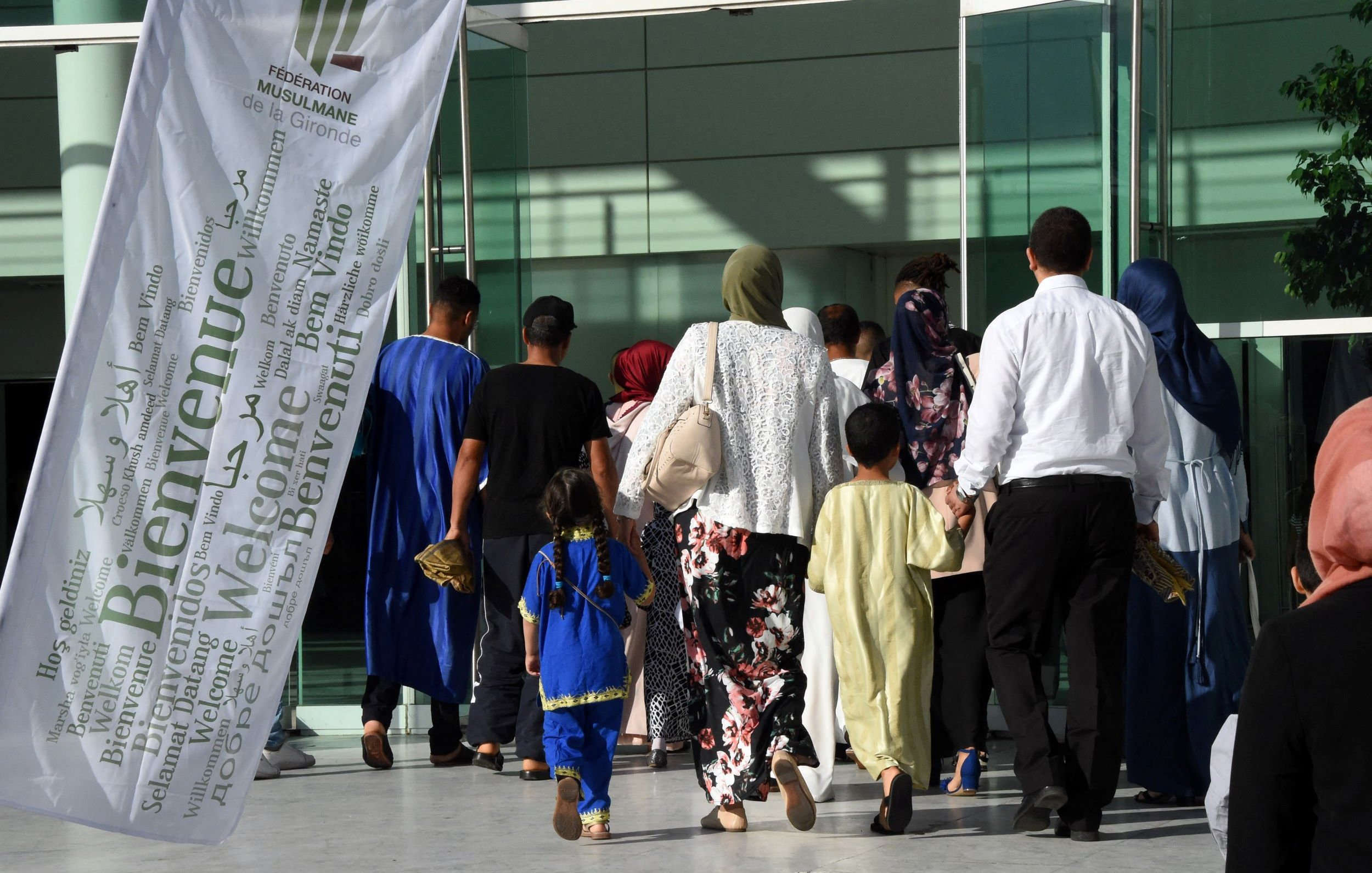 L'image montre un groupe de personnes se dirigeant vers un bâtiment. On peut voir des individus de différentes générations, y compris des enfants. Certains portent des vêtements traditionnels, et plusieurs femmes ont des foulards. Il y a également une grande bannière blanche flottant au vent, portant le mot "Bienvenue" en plusieurs langues. L'ambiance semble chaleureuse et accueillante.