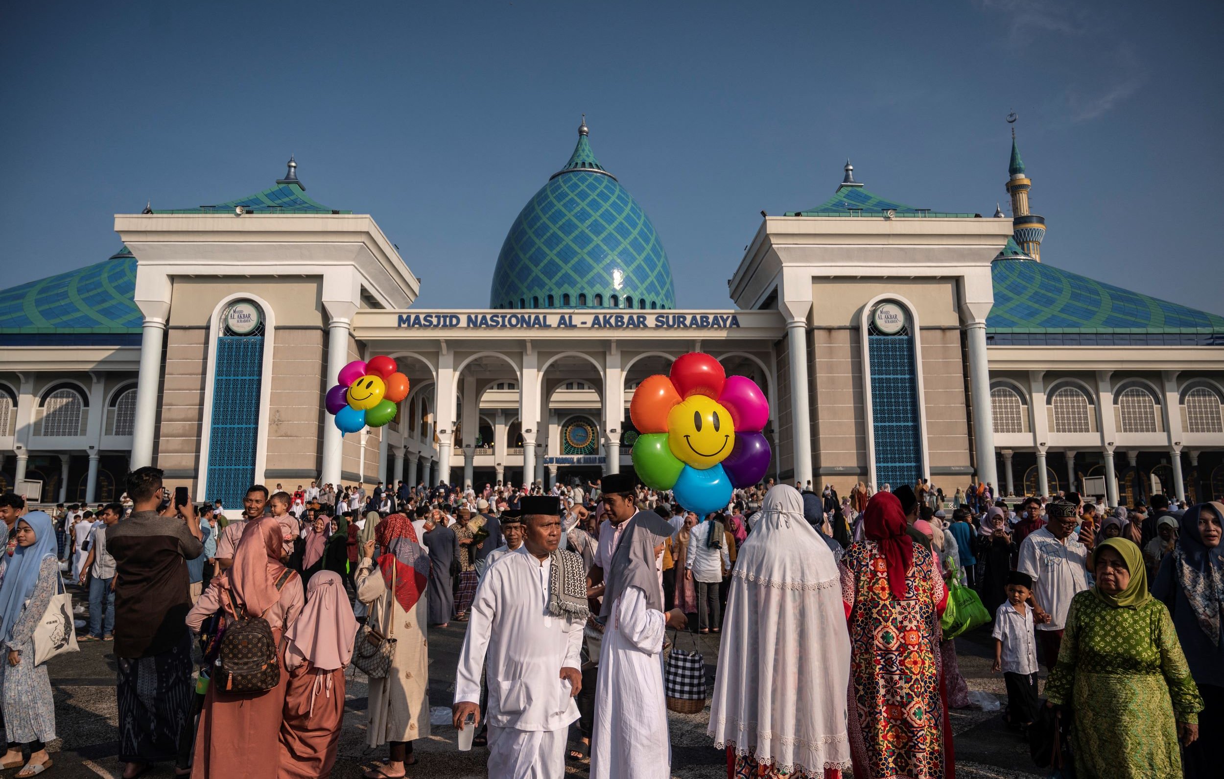 L'image montre un grand rassemblement devant la Masjid Nasional Al-Akbar de Surabaya, en Indonésie. La mosquée est impressionnante, avec des dômes bleu turquoise et une architecture moderne. Beaucoup de personnes sont présentes, certaines portant des vêtements traditionnels, et elles semblent participer à une célébration ou un événement. Des ballons colorés en forme de visages souriants ajoutent une touche festive à la scène. Le ciel est clair, suggérant une belle journée ensoleillée.