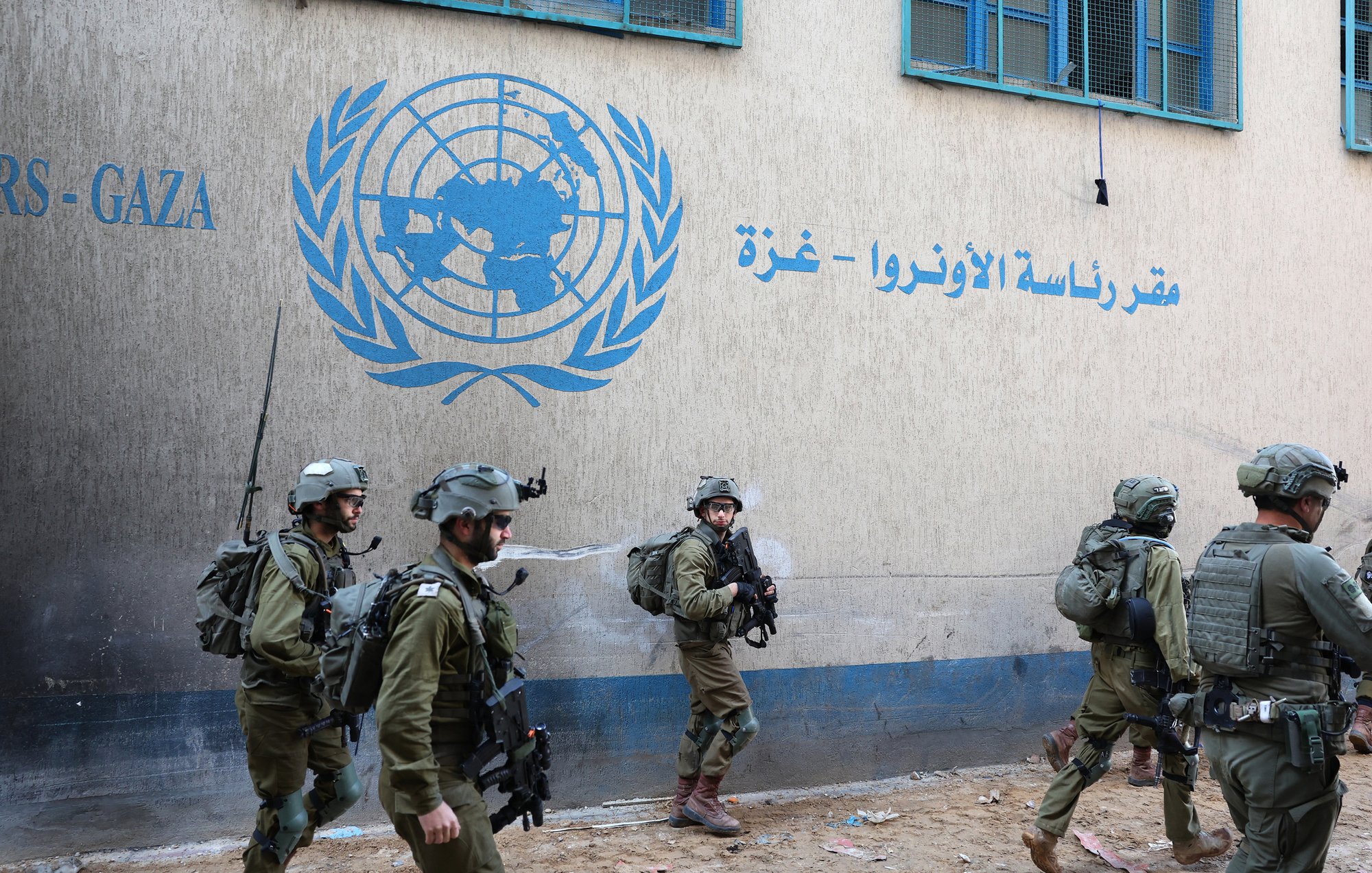 L'immagine mostra un gruppo di soldati in uniforme militare che camminano davanti a un edificio con un logo delle Nazioni Unite, situato nella regione di Gaza. I soldati indossano elmetti e sono equipaggiati per situazioni di operazioni di sicurezza. L'atmosfera è seria e il contesto suggerisce una situazione di tensione o di operazioni militari nella zona. Sul muro dell'edificio si vedono scritte in arabo e in inglese, che indicano che si tratta di un ufficio dell'UNRWA, l'Agenzia delle Nazioni Unite per i Rifugiati Palestinesi.